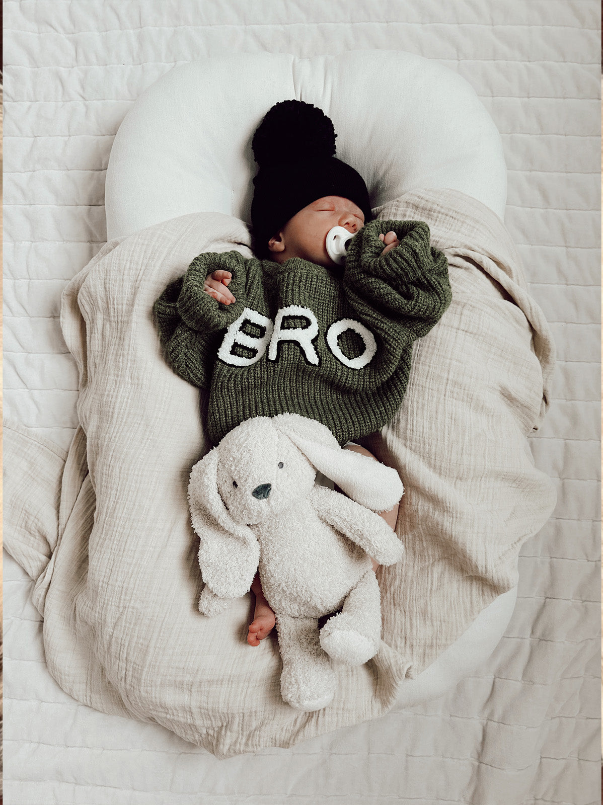 Infant in green sweater with "BRO" resting on a blanket, holding a white plush bunny. Cozy indoor setting.