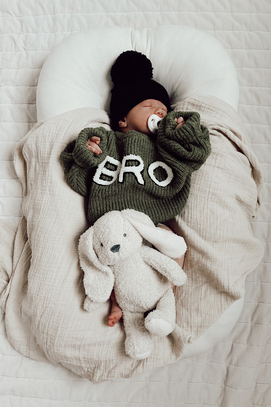 Infant in green sweater with "BRO" resting on a blanket, holding a white plush bunny. Cozy indoor setting.