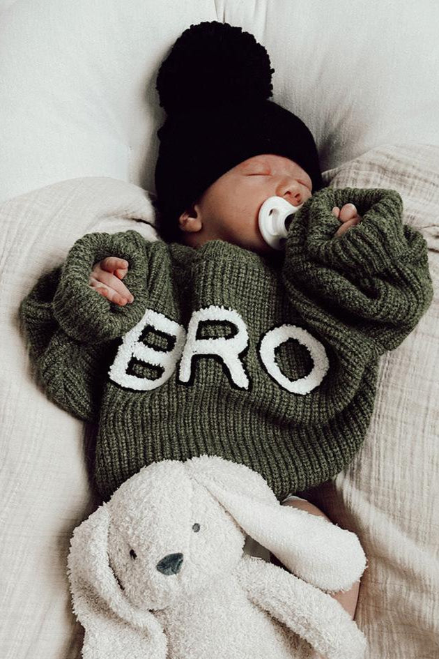 Baby wearing a green "BRO" sweater and black hat, cuddling a soft bunny toy while sleeping peacefully.