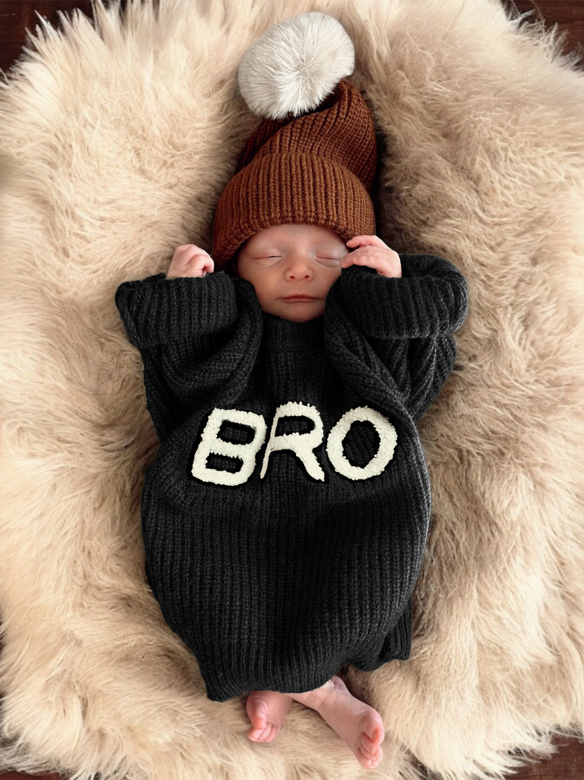 Newborn baby sleeping on a fluffy rug, wearing a black sweater with "BRO" and a brown hat with a pompom.