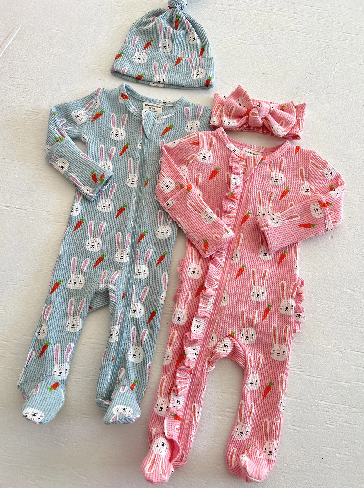 Two bunny-patterned baby onesies and matching hat and headband, in blue and pink, displayed on a white surface.