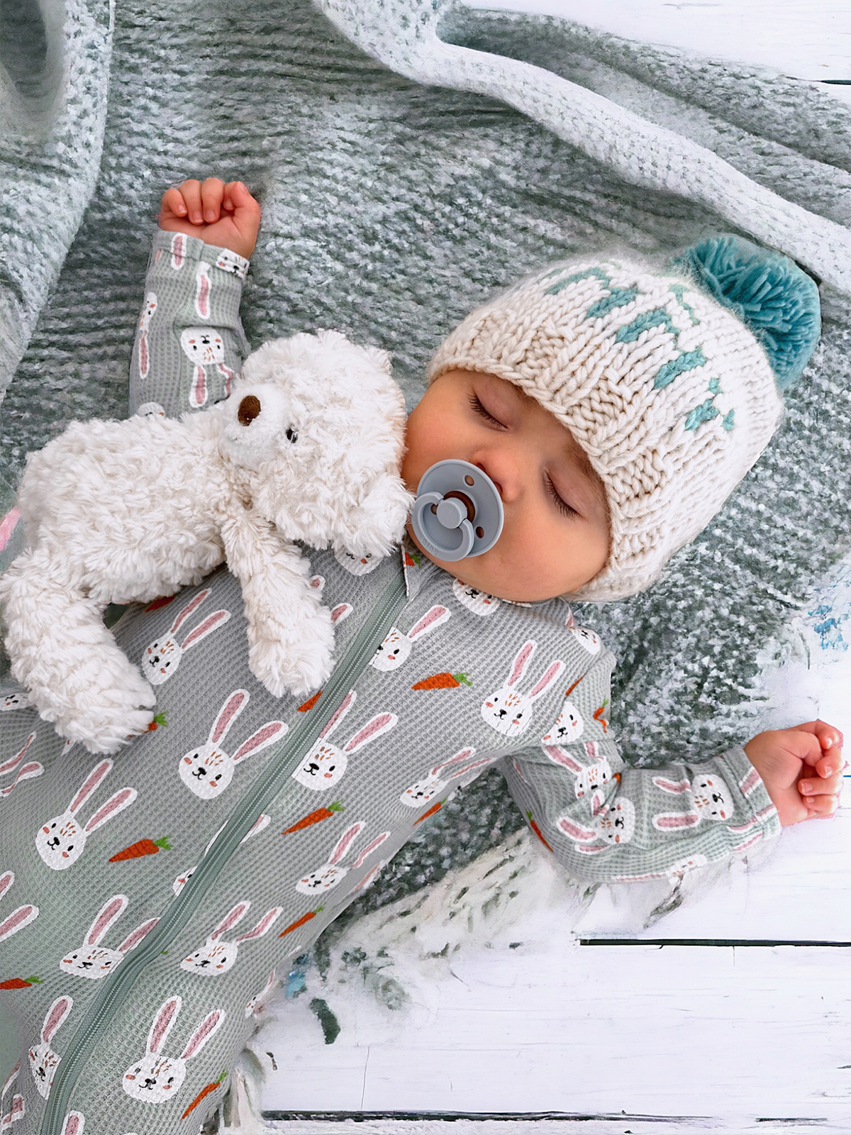 Baby sleeping peacefully in a bunny-patterned onesie, holding a plush bear and wearing a knitted hat on a cozy blanket.