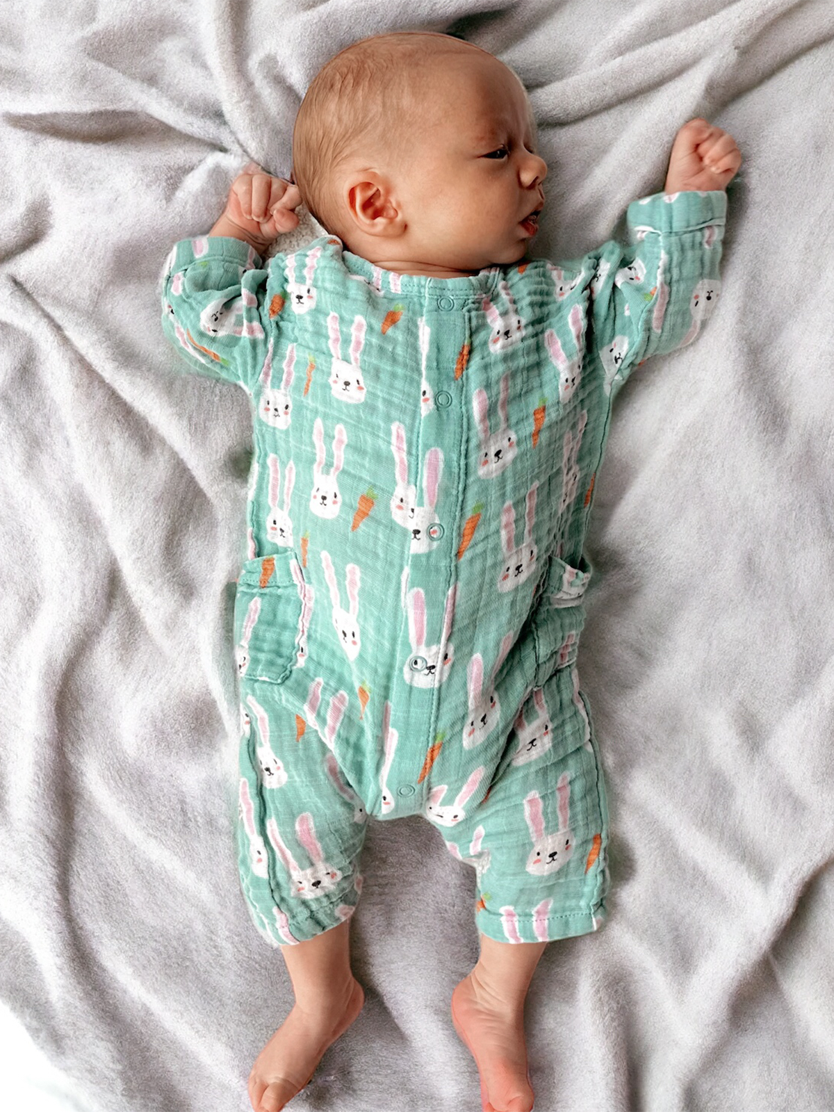 Baby in a green bunny-patterned outfit lying on a soft gray blanket, arms raised in a relaxed pose.