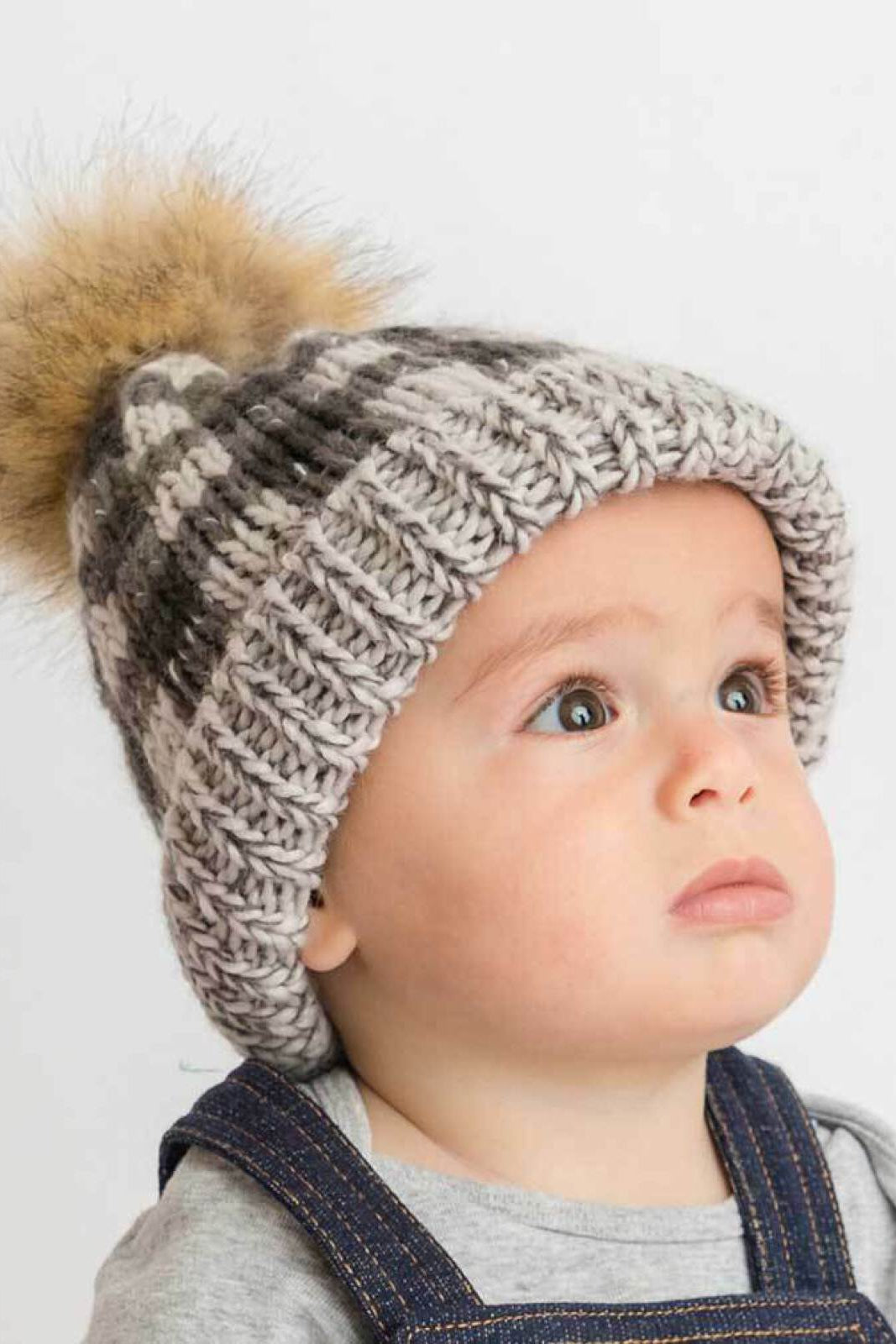 Baby wearing a knitted gray and white striped beanie with a pom-pom, looking up with a thoughtful expression.