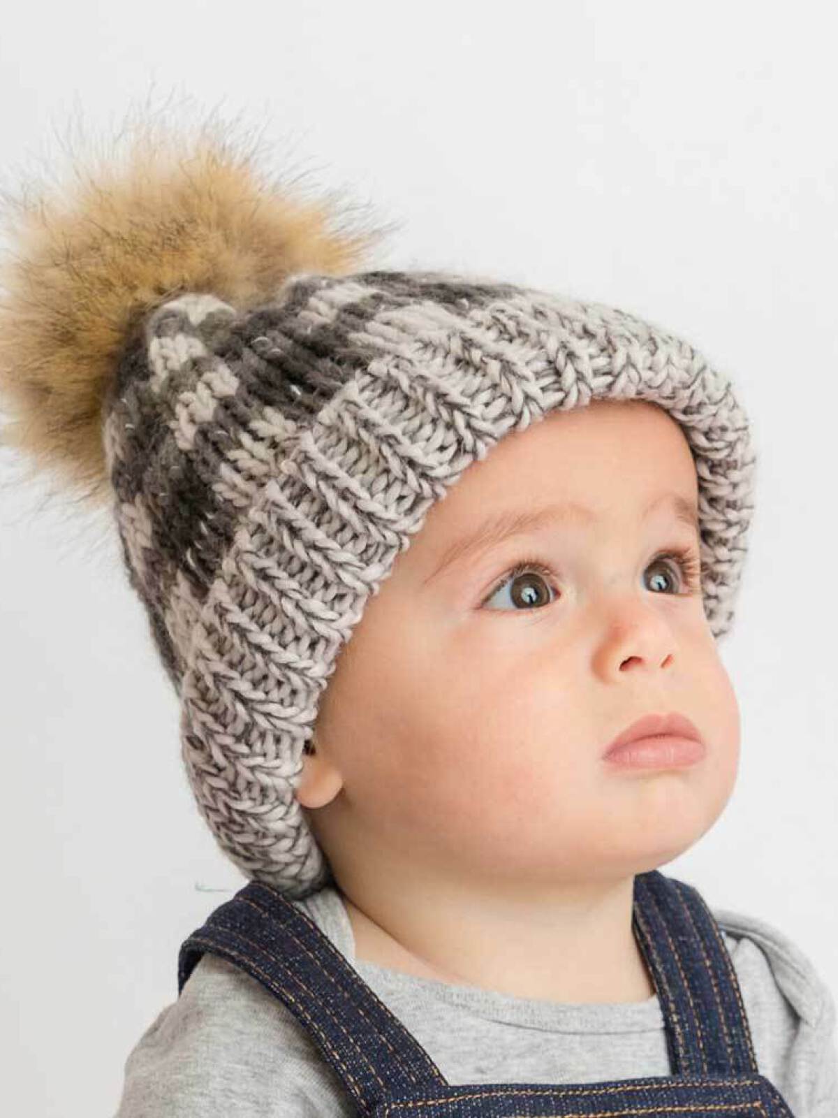 Baby wearing a knitted gray and white striped beanie with a pom-pom, looking up with a thoughtful expression.