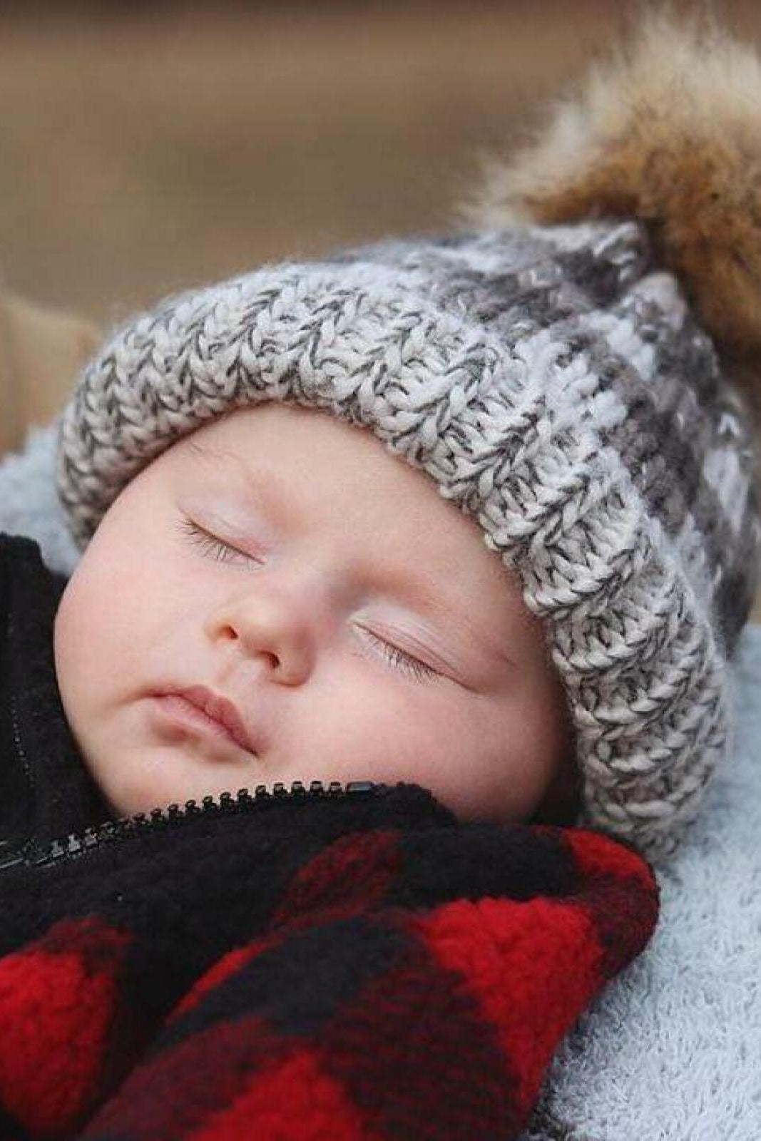 Sleeping baby in a cozy knitted hat and red plaid blanket, peacefully resting outdoors.