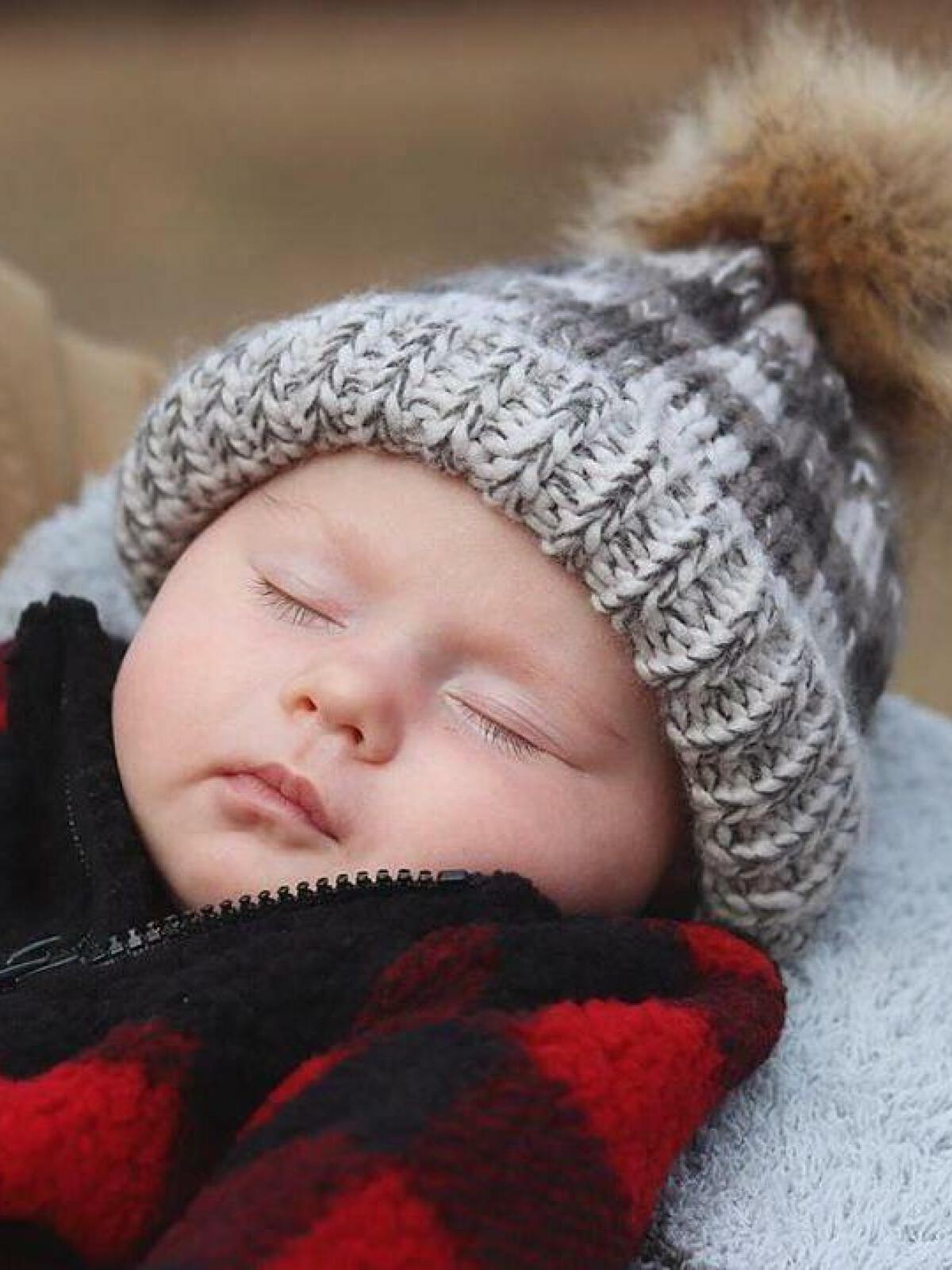Sleeping baby in a cozy knitted hat and red plaid blanket, peacefully resting outdoors.