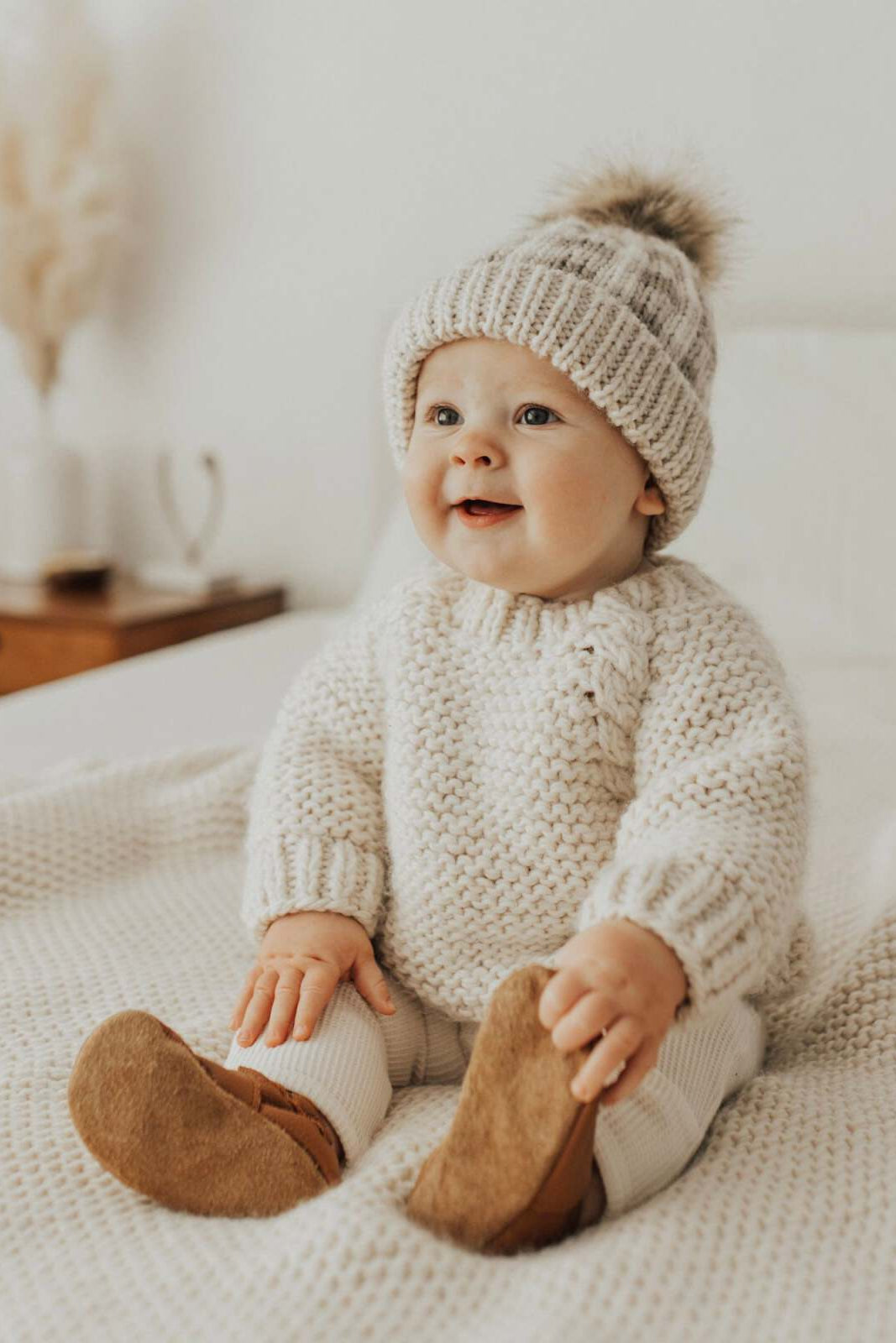 Smiling baby in a cozy knitted sweater and hat, seated on a soft blanket in a bright room.
