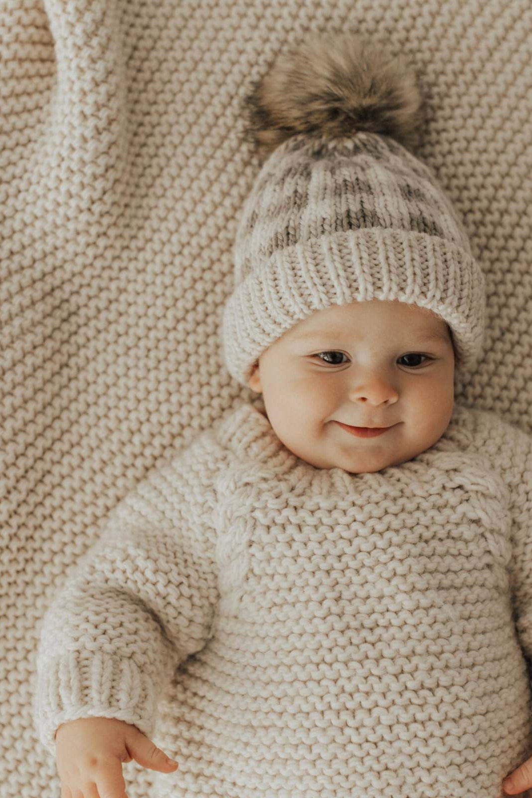 Smiling baby in a knitted sweater and hat on a textured blanket, creating a warm, cozy scene.