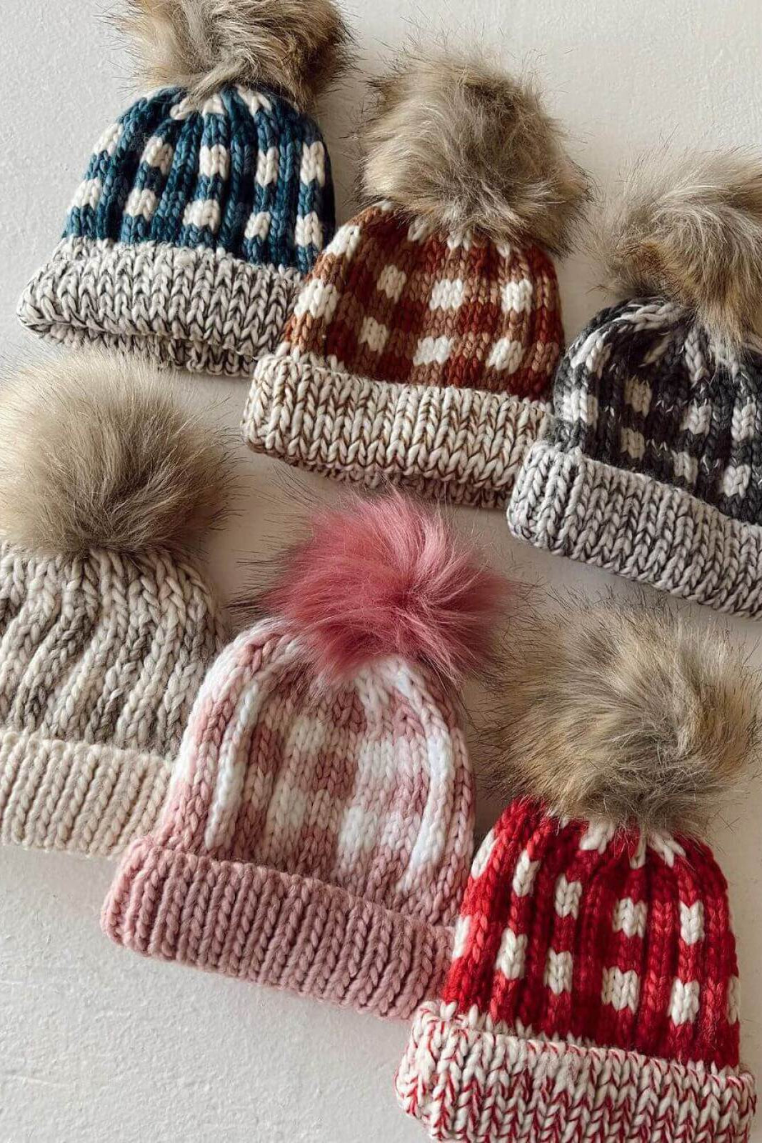 Six knitted hats with fur pom-poms in various colors and patterns laid out on a light background.