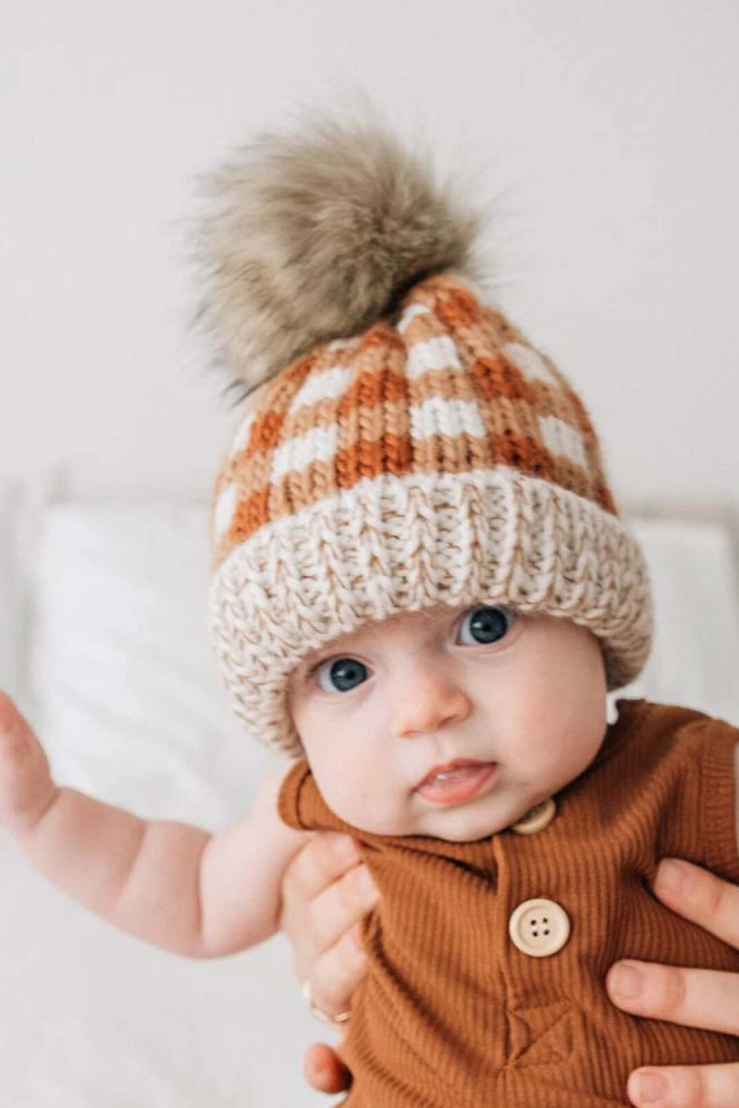 Baby in a knitted hat with a pom-pom, wearing a brown onesie, held by a person against a light background.