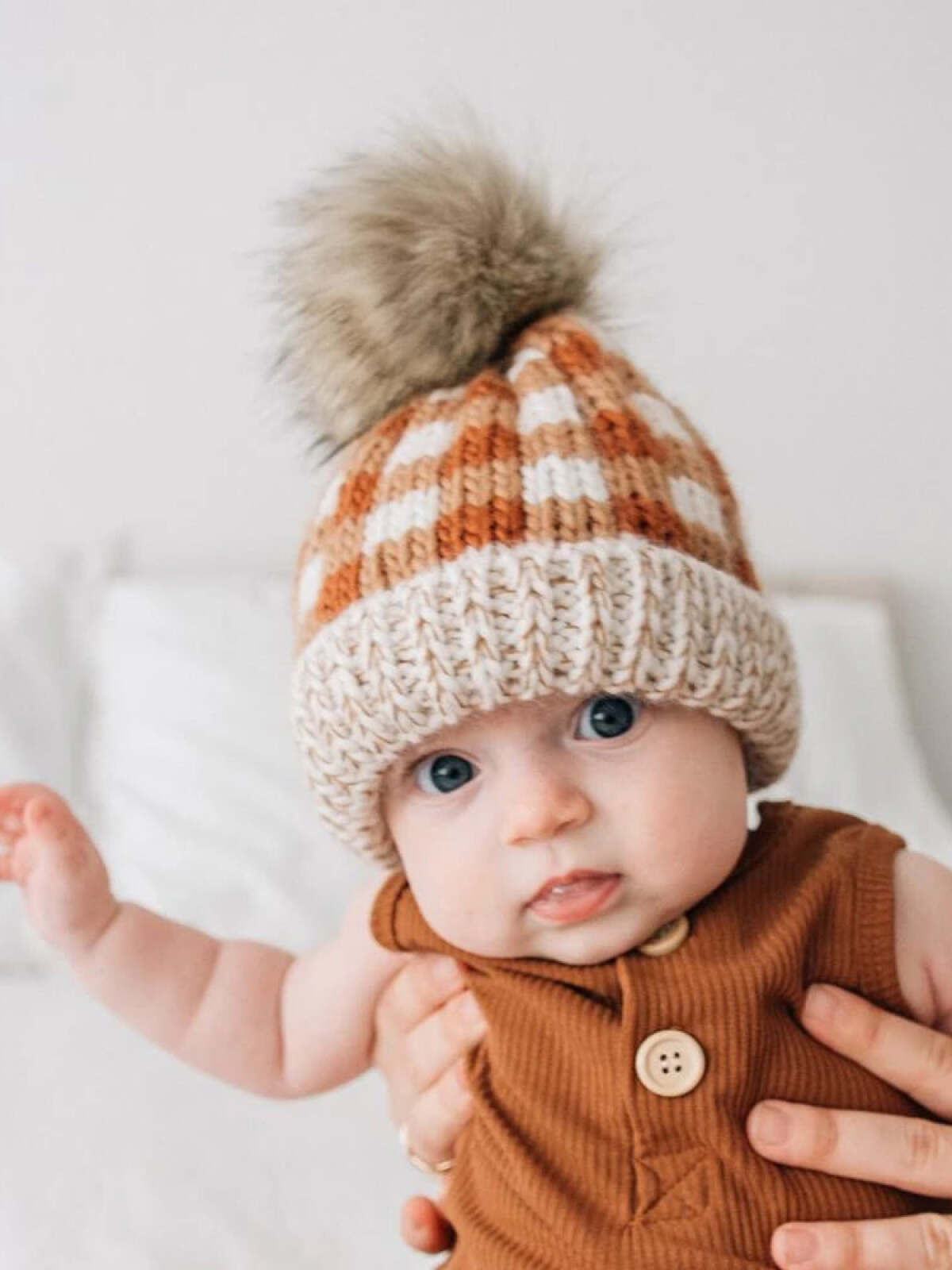 Baby in a knitted hat with a pom-pom, wearing a brown onesie, held by a person against a light background.