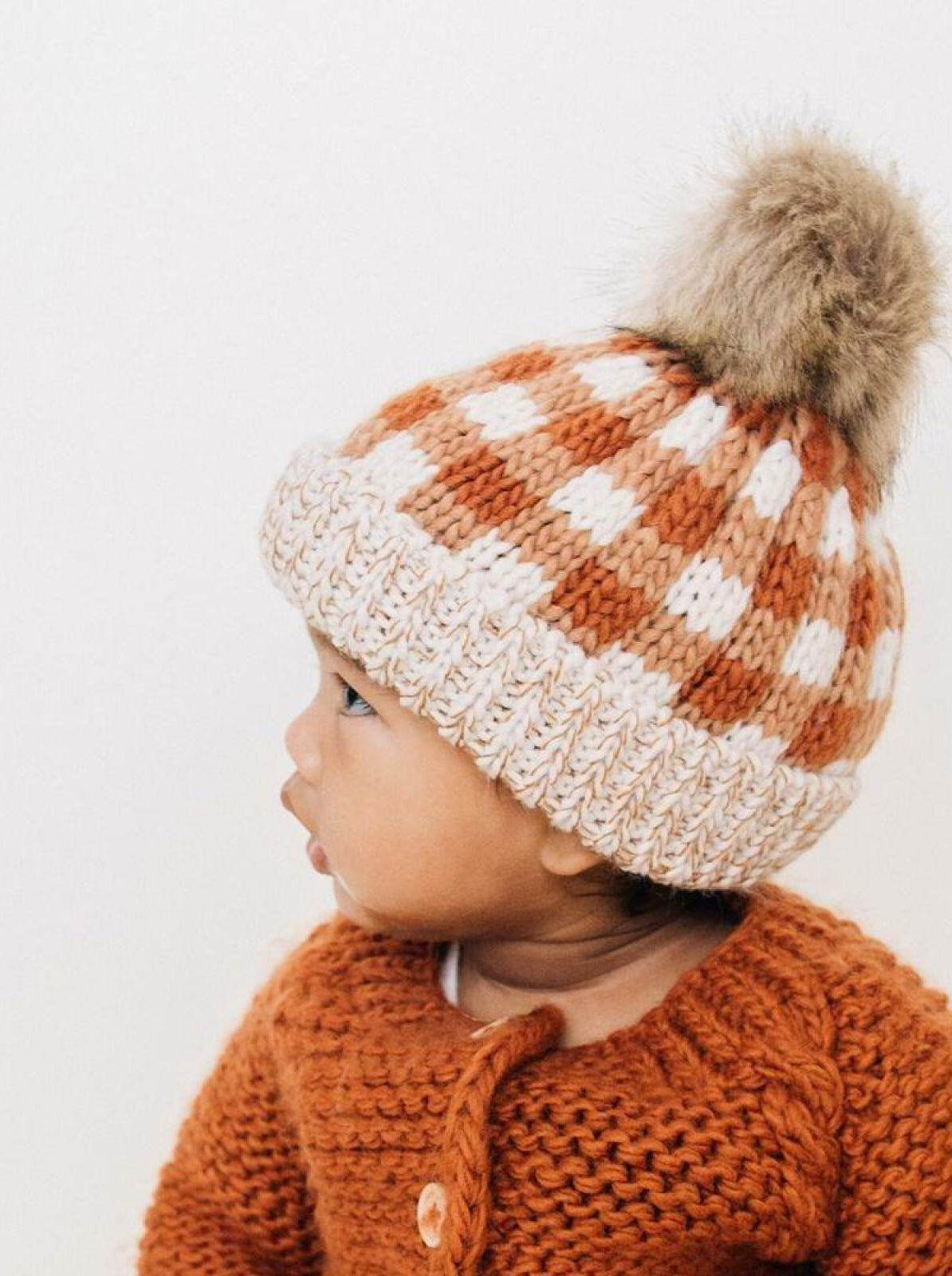 Baby wearing a knitted checkered hat with a pom-pom, dressed in an orange sweater.