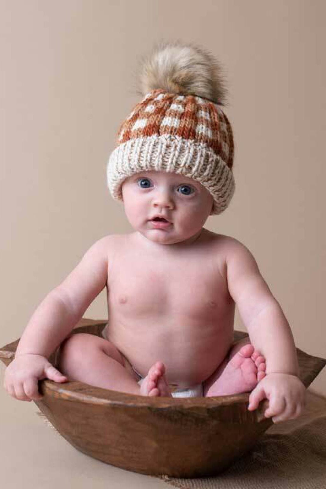 Baby sitting in a wooden bowl, wearing a knitted hat with a pom-pom, against a beige background.