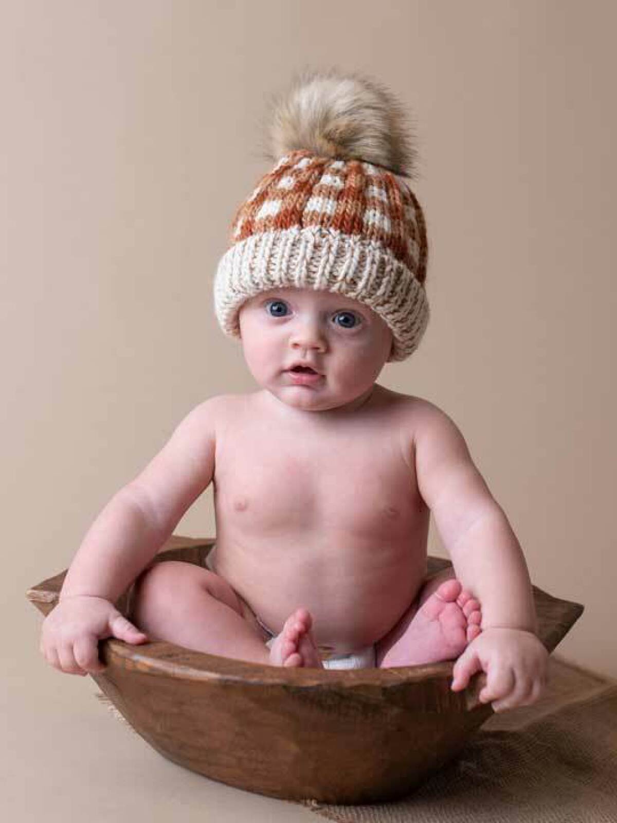 Baby sitting in a wooden bowl, wearing a knitted hat with a pom-pom, against a beige background.