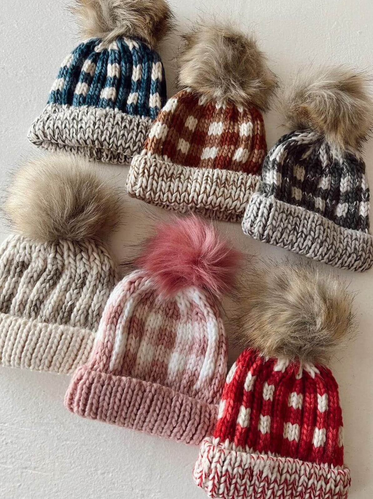 Six knitted beanies with fur pom-poms in various colors and patterns, arranged on a light surface.