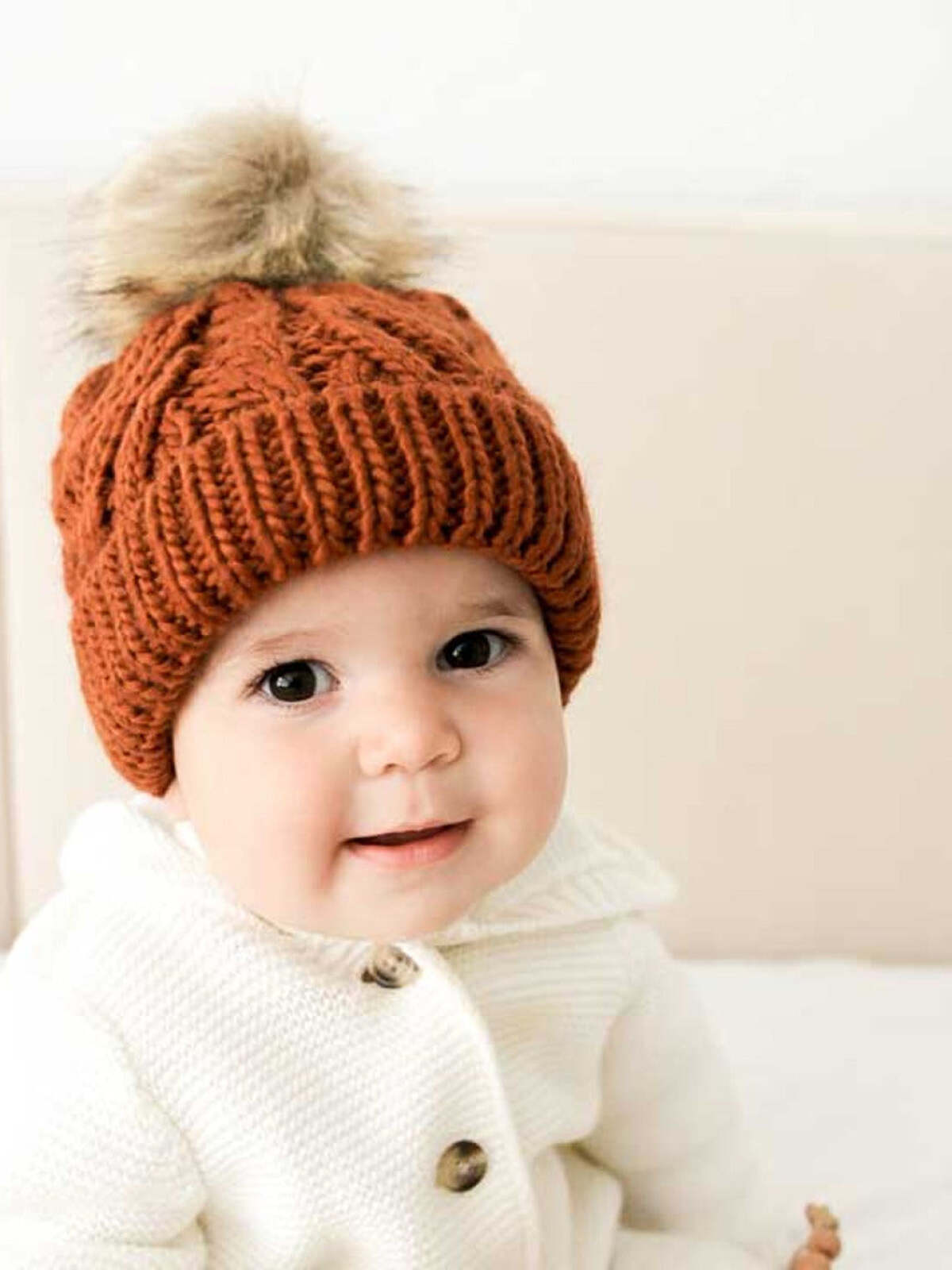 Baby smiling while wearing a cozy orange knitted hat with a pom-pom, dressed in a white sweater.