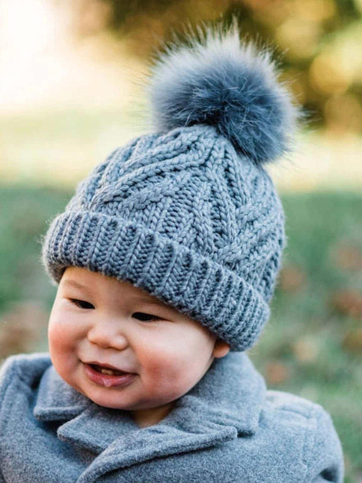 Smiling baby in a gray knitted hat with a pom-pom, wearing a gray coat outdoors.