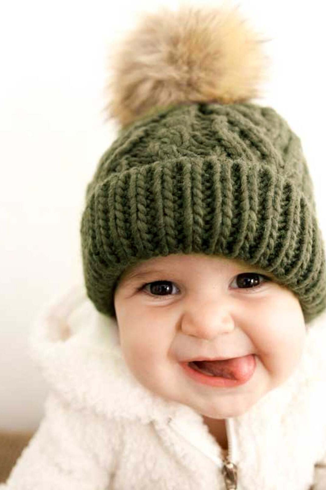 Smiling baby wearing an olive green knit hat with a pom-pom, sticking out tongue, and cozy white jacket.