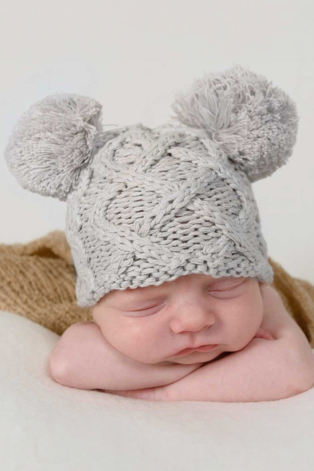 Newborn baby sleeping peacefully, wearing a cozy gray knitted hat with pom-poms. Soft textures and calm setting.