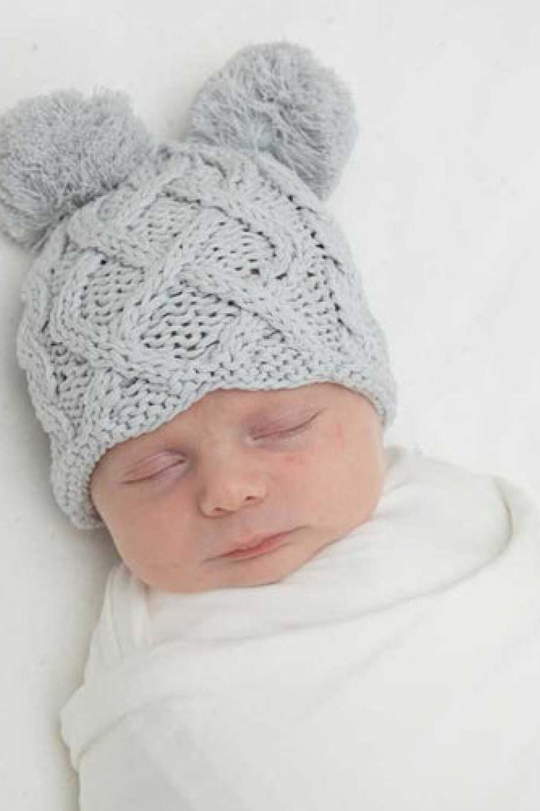 Baby sleeping peacefully, wearing a light gray knitted hat with pom-poms, wrapped in a white blanket.