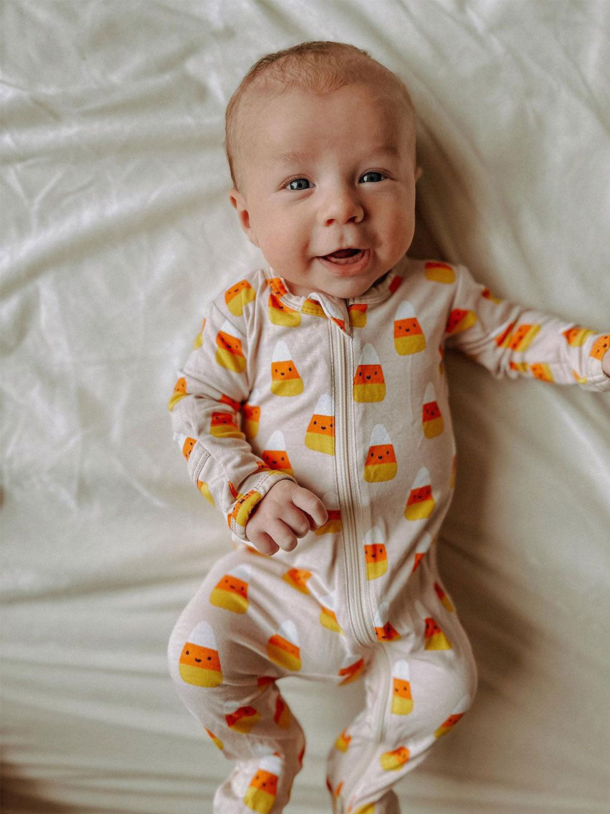 Smiling baby in candy corn-patterned pajamas lying on a soft, white blanket.