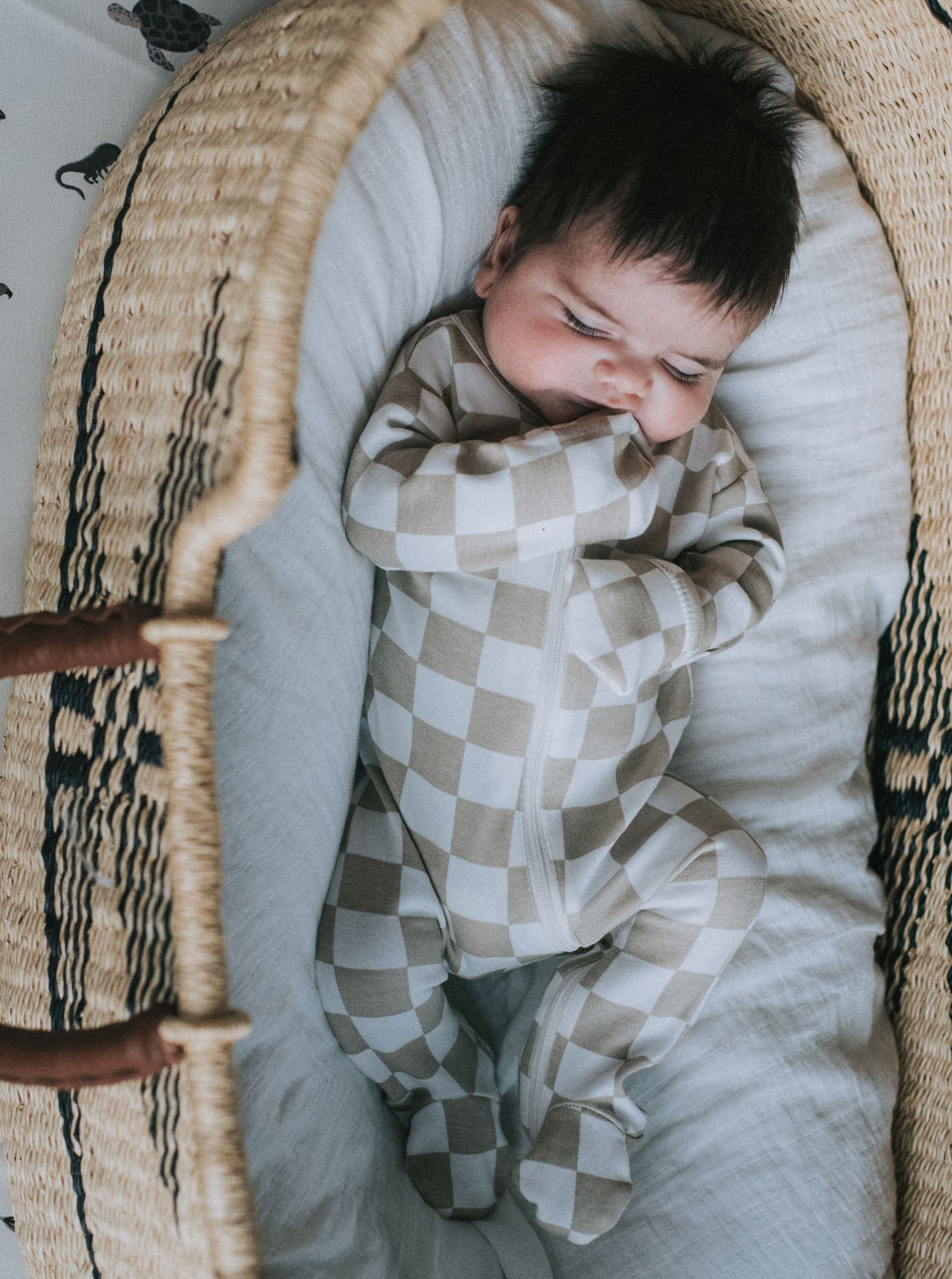 Infant in a checkered onesie lies peacefully in a woven bassinet, surrounded by soft white fabric.