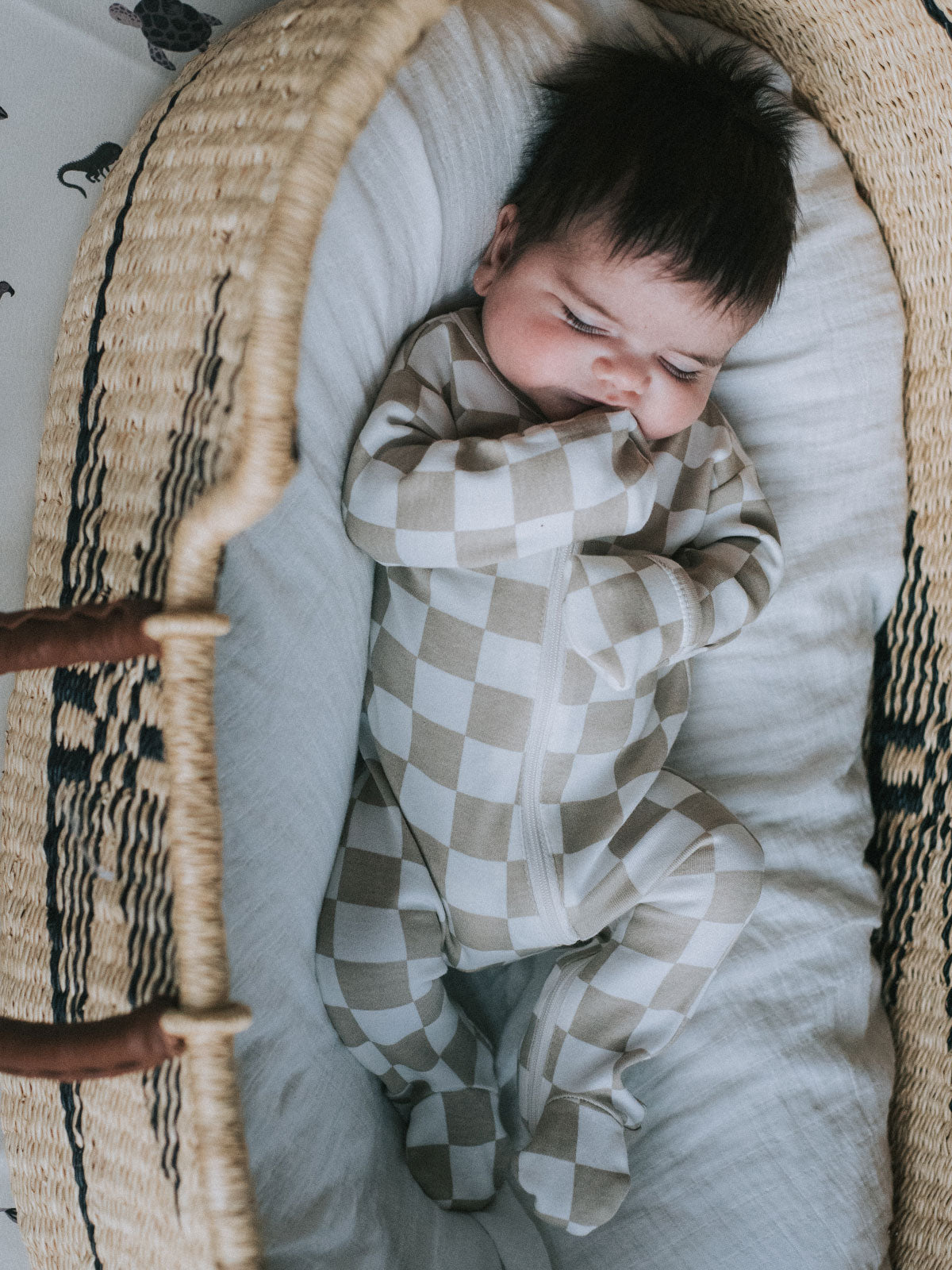 Infant in a checkered onesie lies peacefully in a woven bassinet, surrounded by soft white fabric.