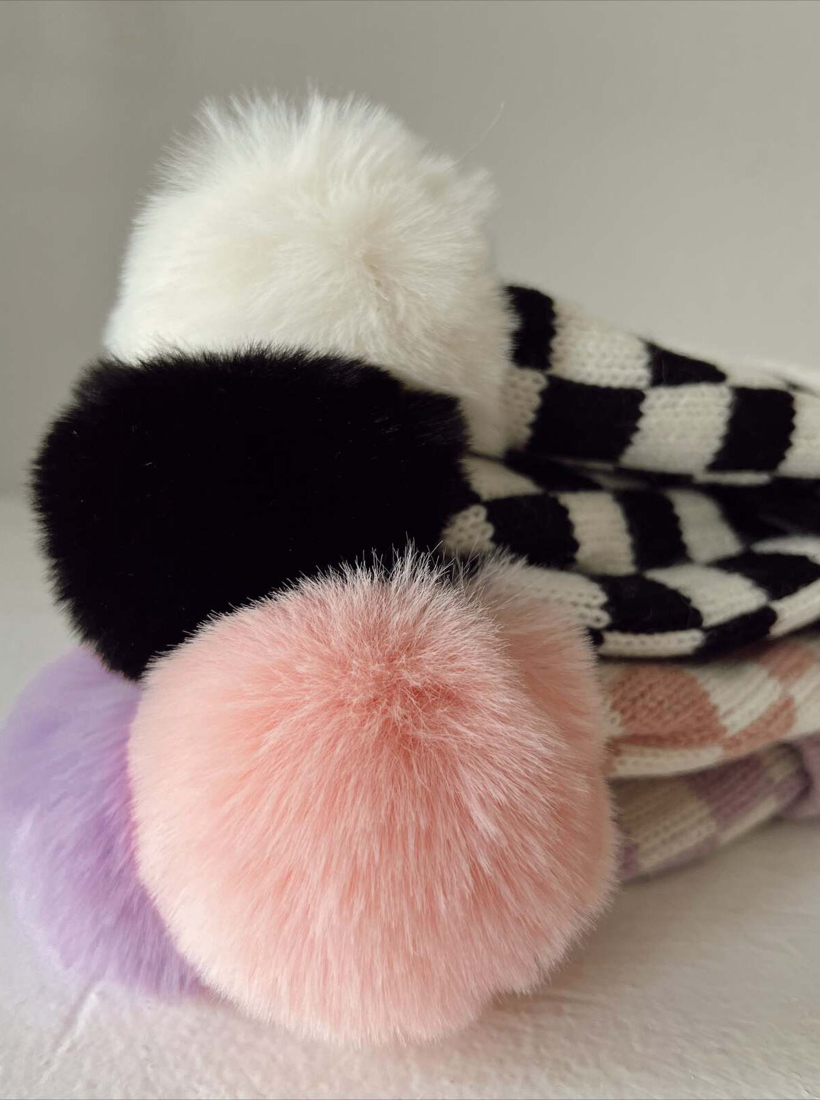 Colorful pom-poms on knit hats stacked together, showcasing white, black, pink, and purple fur textures.