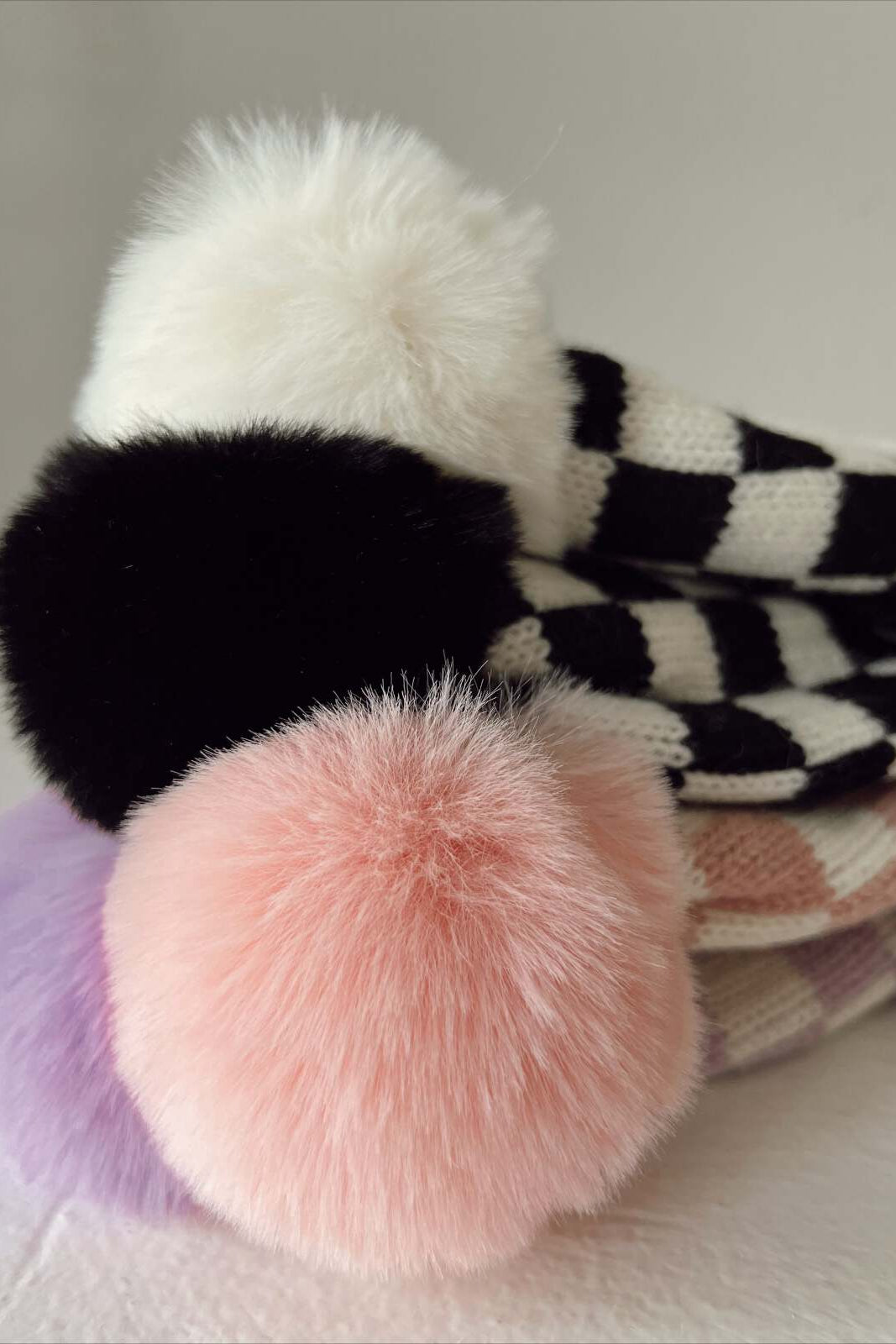 Colorful pom-poms on knit hats stacked together, showcasing white, black, pink, and purple fur textures.