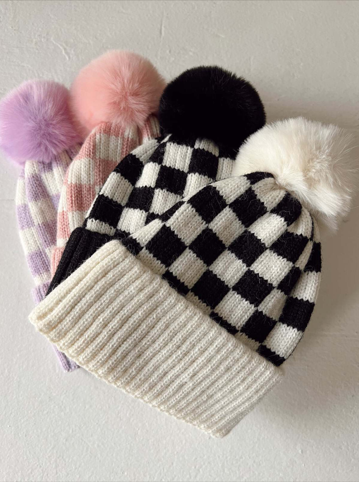 Four stylish knit beanie hats with pom-poms in black, white, pink, and purple patterns, displayed on a light surface.