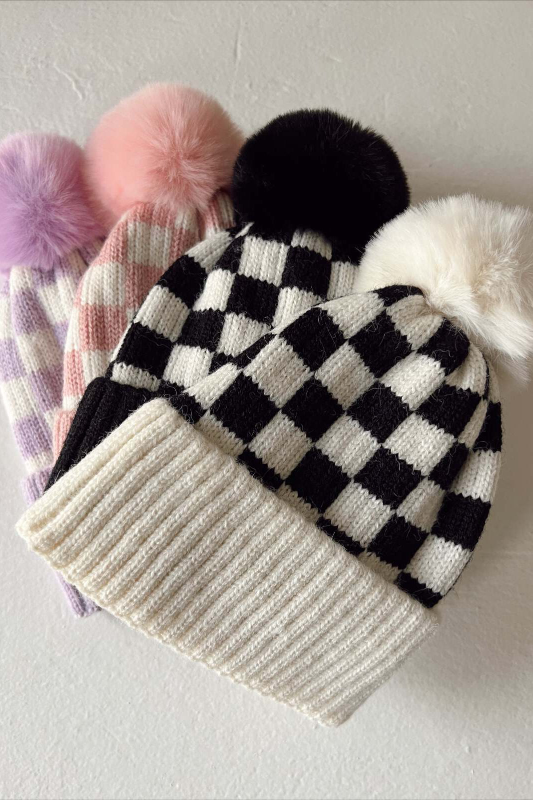 Four stylish knit beanie hats with pom-poms in black, white, pink, and purple patterns, displayed on a light surface.
