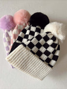 Four stylish knit beanie hats with pom-poms in black, white, pink, and purple patterns, displayed on a light surface.