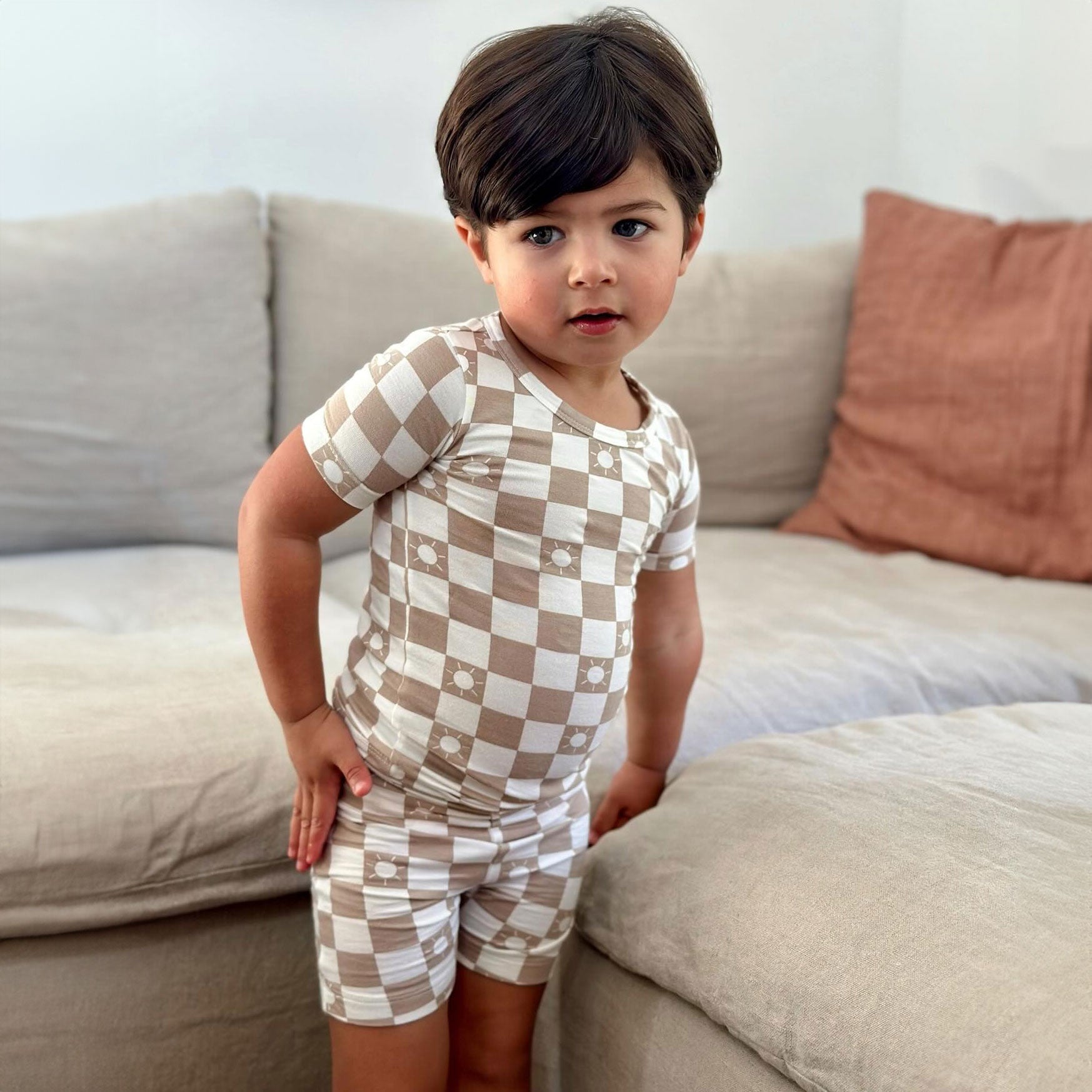 Toddler in checkered beige outfit poses on a sofa with neutral decor.