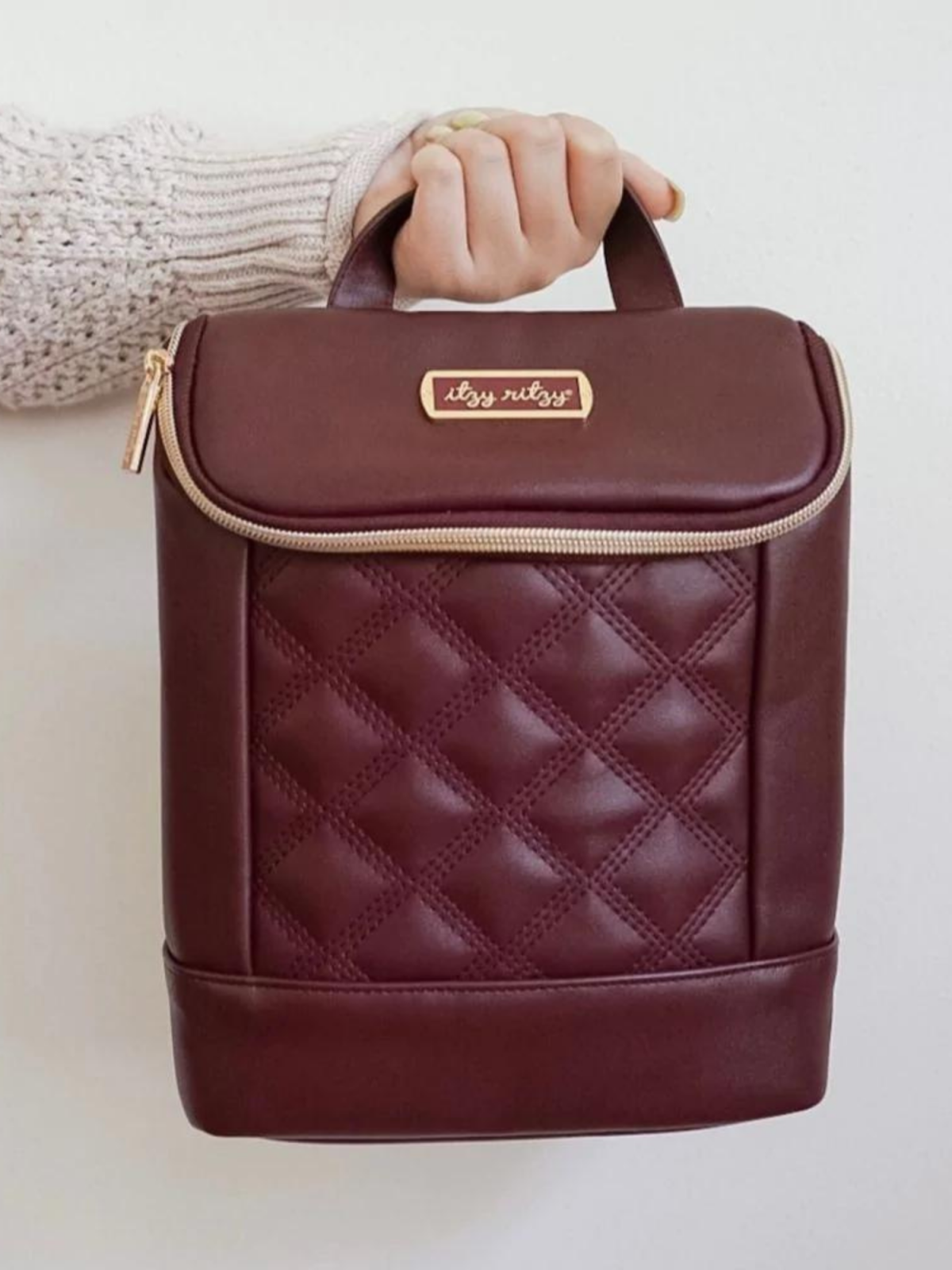 Burgundy quilted backpack with a gold logo, held by a hand in a cozy sweater.