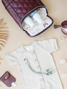 Beige baby onesie with buttons, pacifier clip, and a burgundy bag with baby bottles inside on a neutral background.