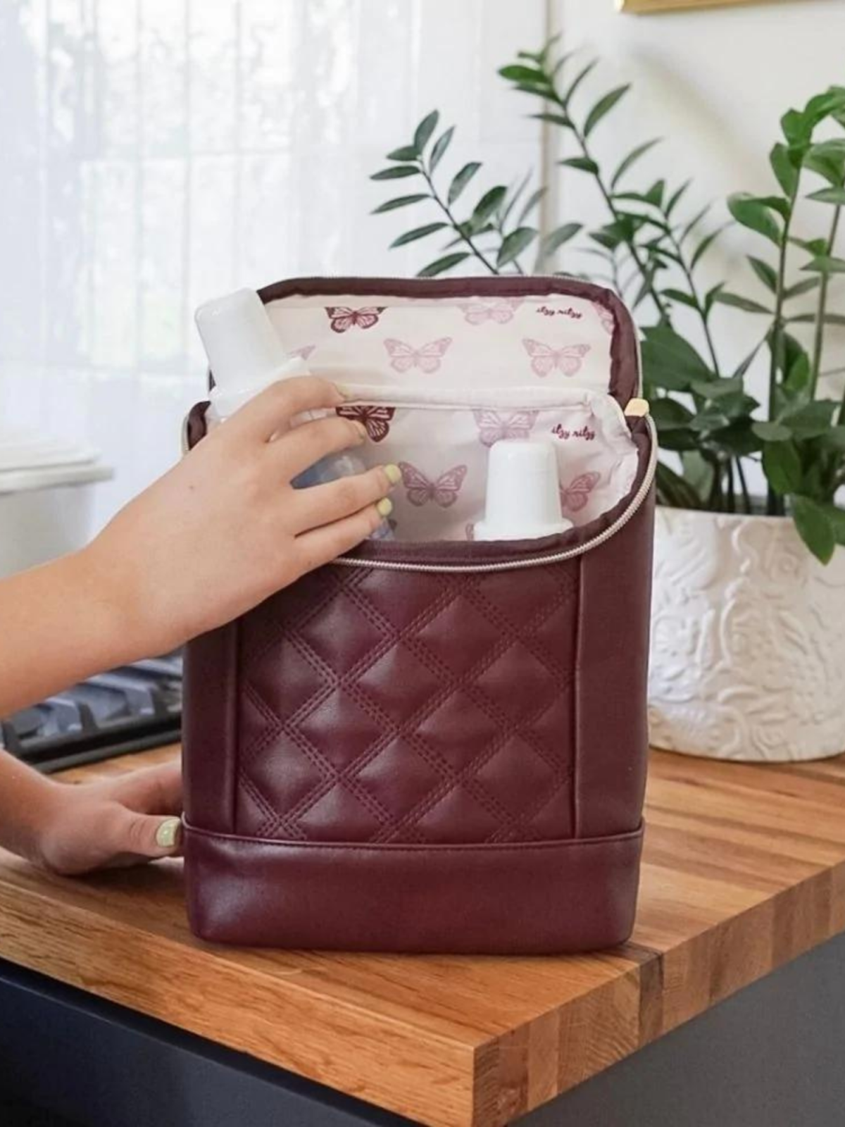 Hand reaching into a burgundy bag with butterfly-patterned lining, set on a wooden table with greenery in the background.