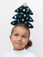 Girl wearing a festive green Christmas tree headband with decorations, looking up with a playful expression.
