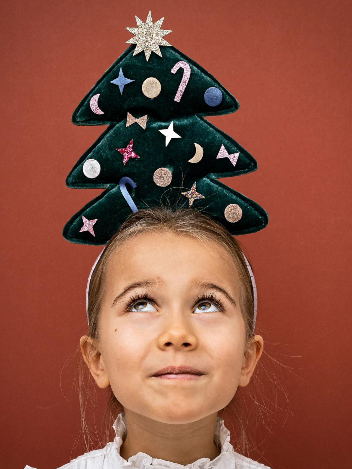 Girl with a Christmas tree hat, looking upwards against a red background, festive decorations on the hat.