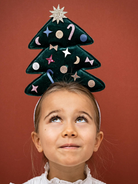 Girl with a Christmas tree hat, looking upwards against a red background, festive decorations on the hat.