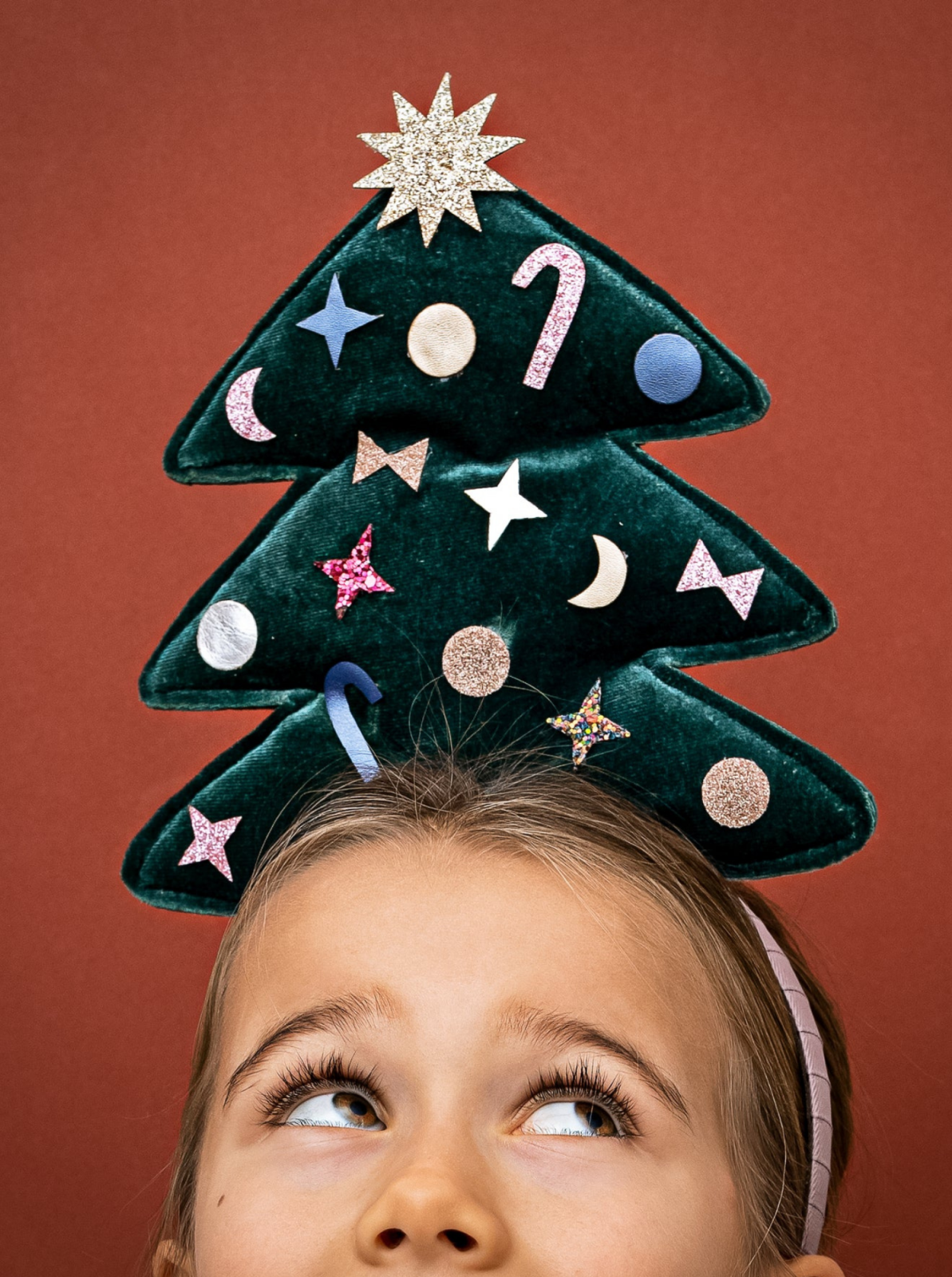 Child wearing a festive Christmas tree headband with colorful decorations, looking up against a reddish background.