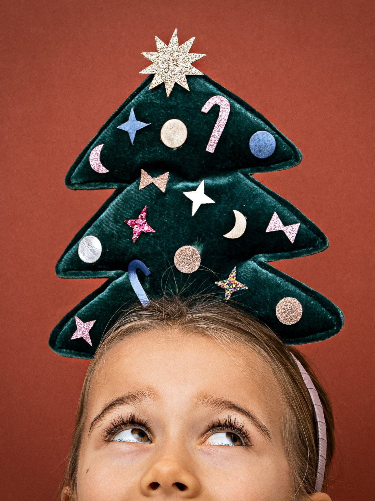 Child wearing a festive Christmas tree headband with colorful decorations, looking up against a reddish background.