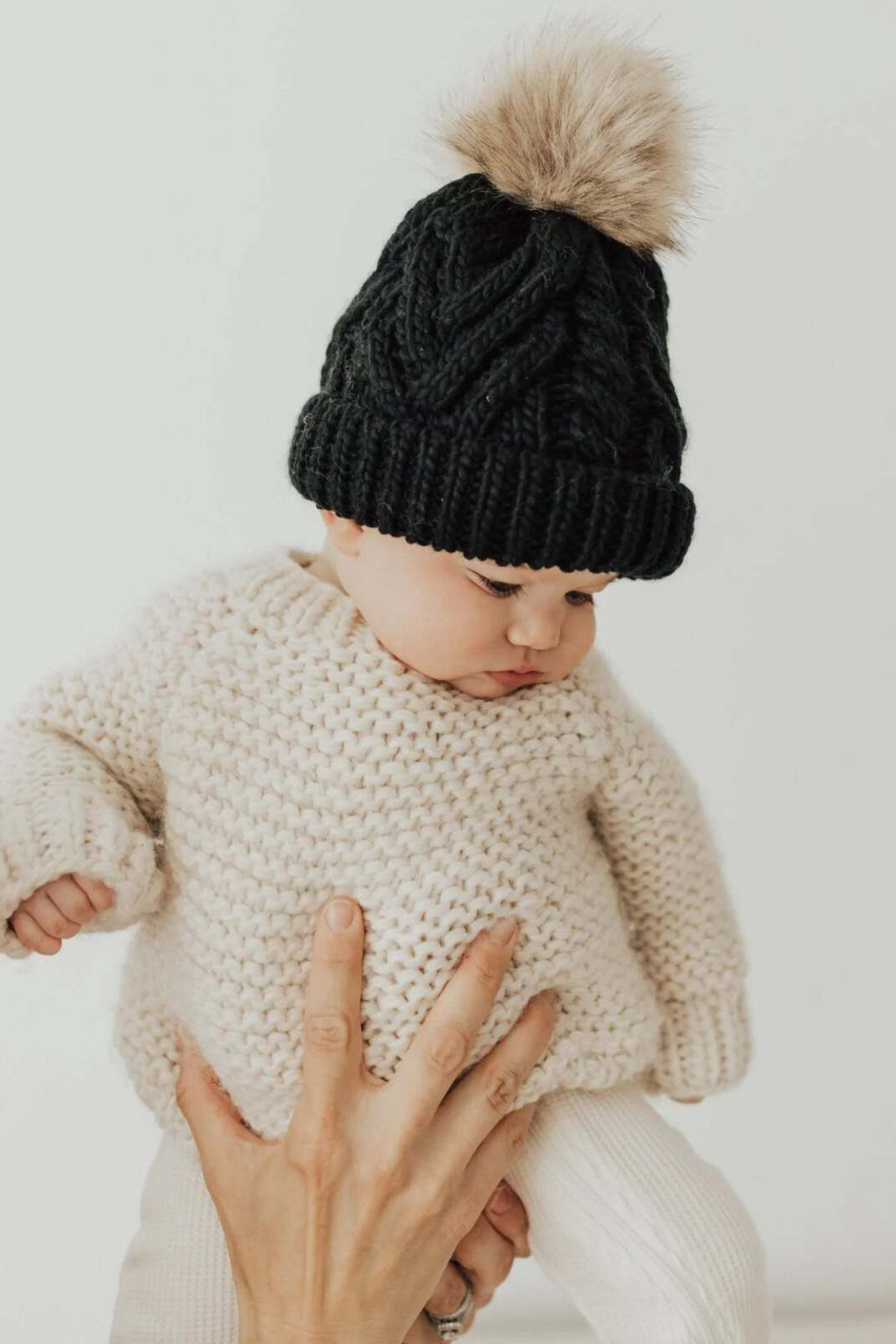 Baby in a cozy cream sweater and black knitted hat with a pom-pom, held by an adult hand.