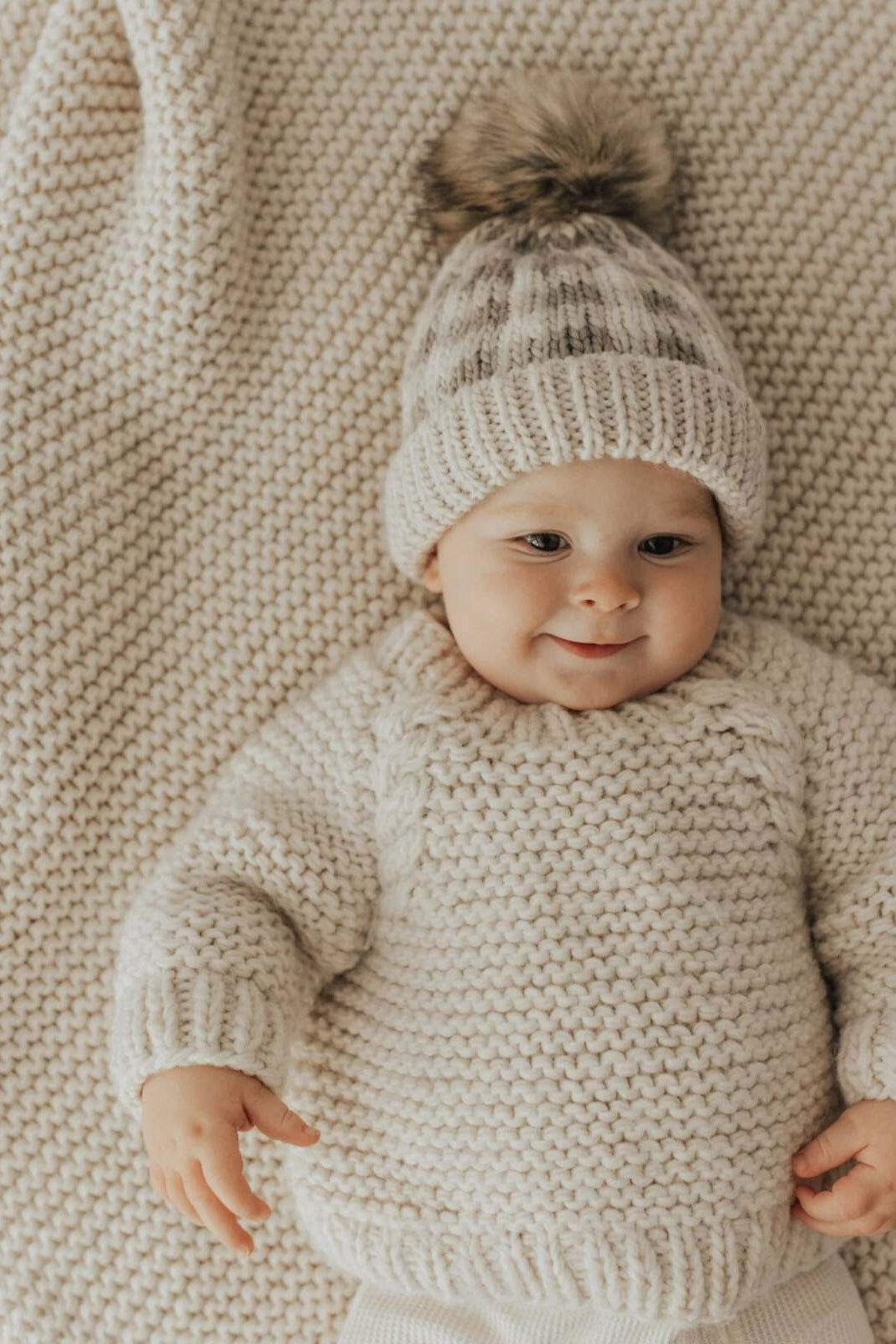 Baby in a cozy knit sweater and hat smiles on a textured blanket. Soft, neutral colors create a warm ambiance.