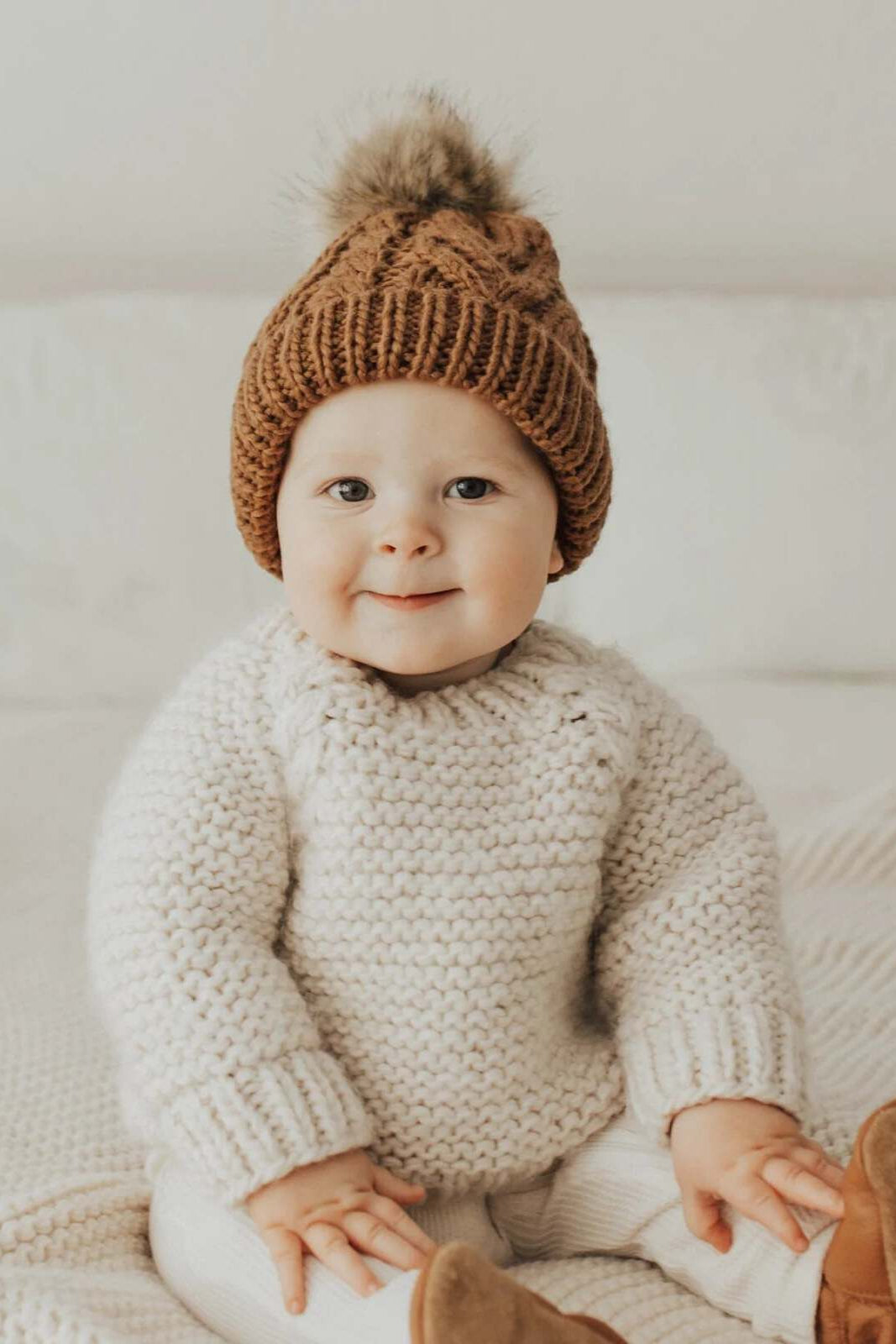 Baby wearing a knitted sweater and brown hat, smiling while sitting on a textured blanket.