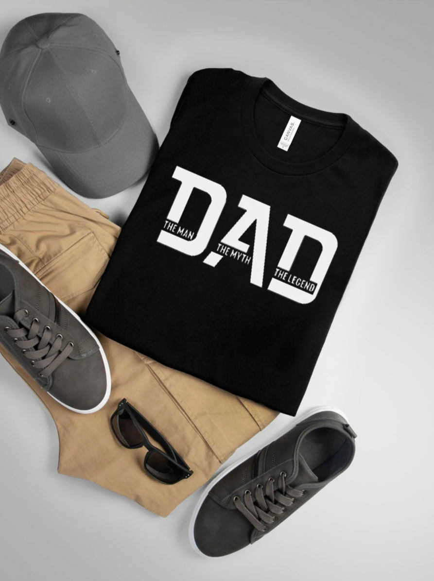 Black t-shirt with "DAD: The Man, The Myth, The Legend" design, paired with khaki shorts, gray shoes, and a cap.