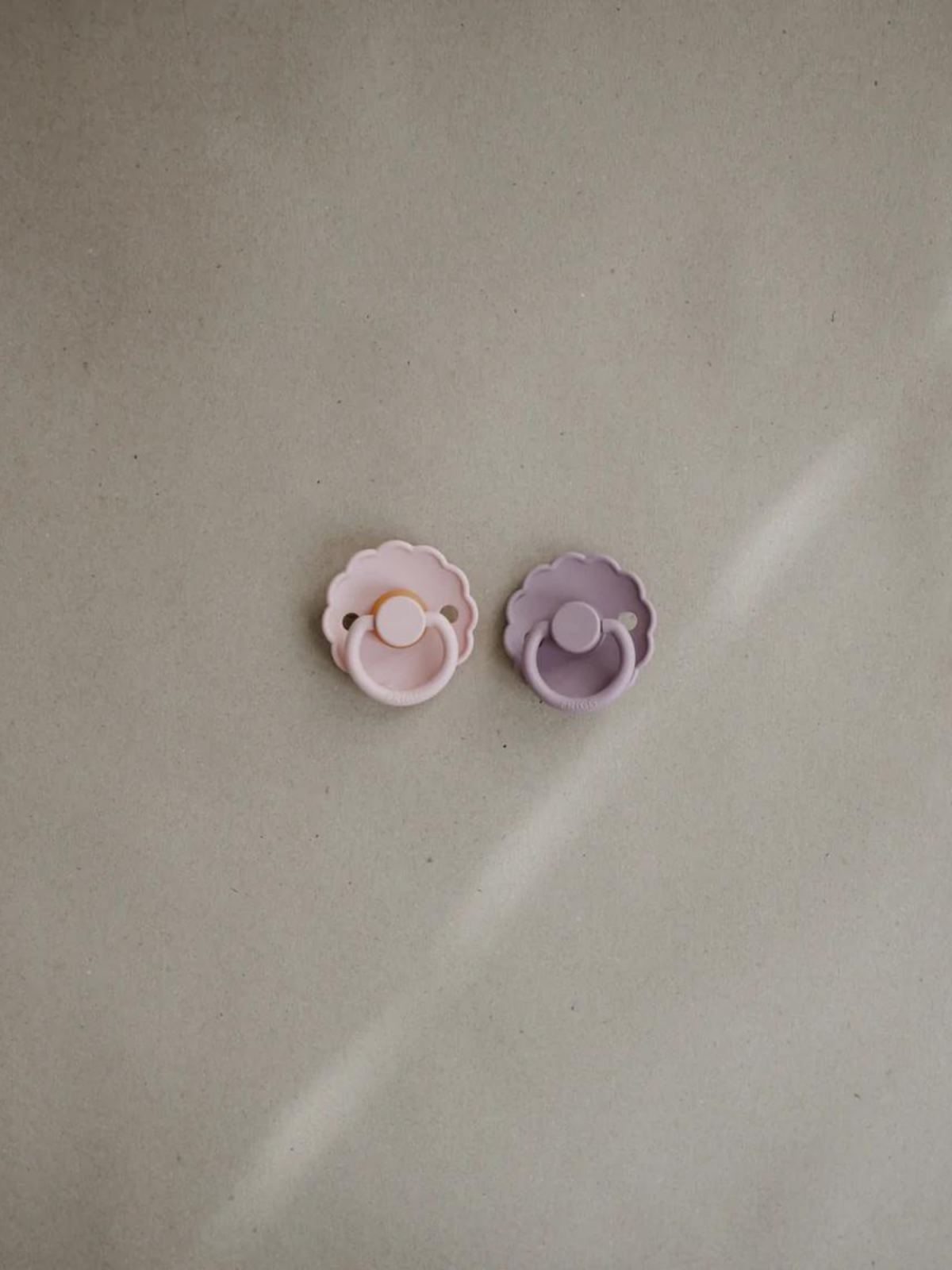 Two silicone pacifiers in pink and purple on a beige background.