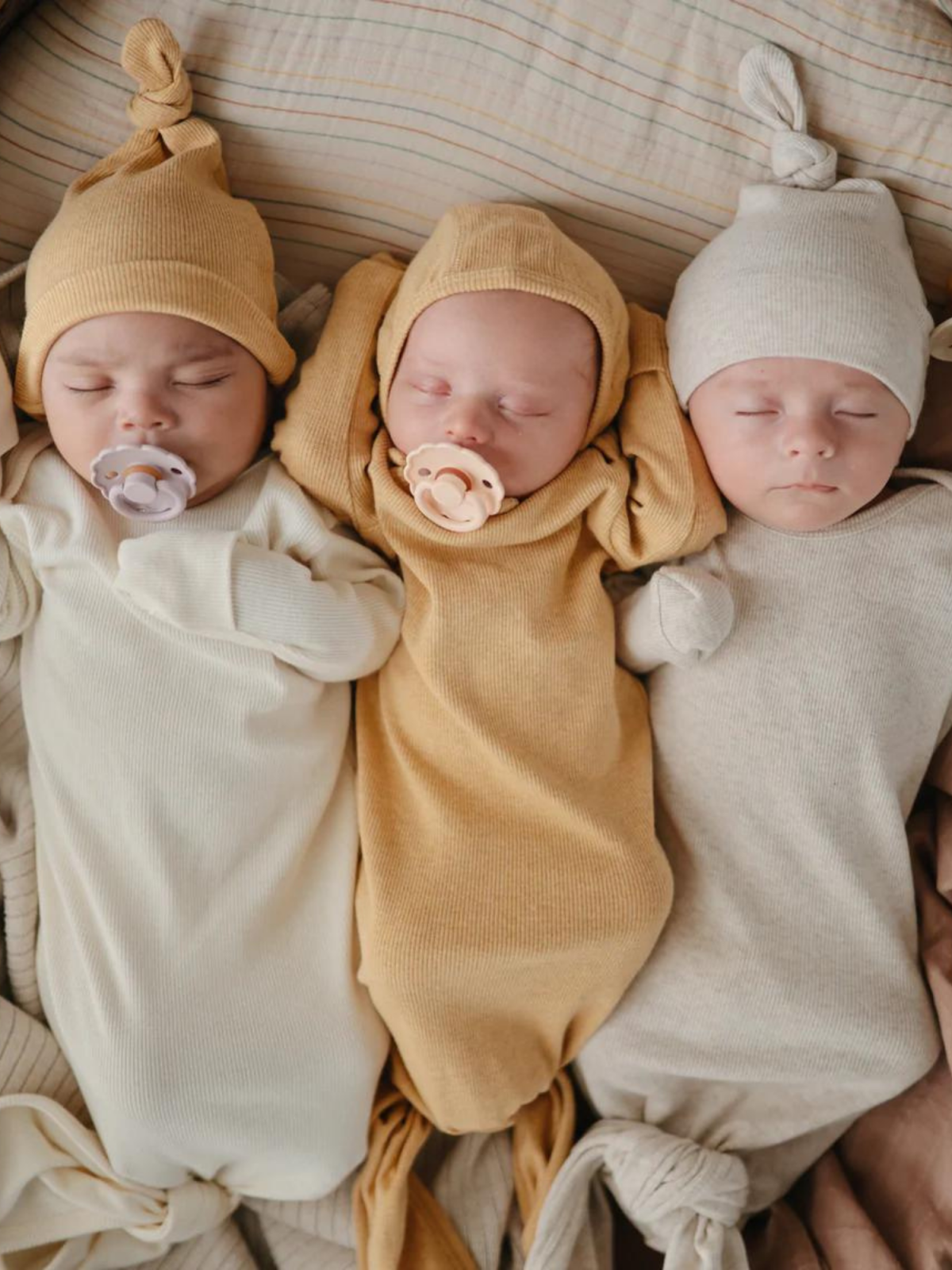 Three sleeping babies in knitted outfits and hats, swaddled in soft fabric on a cozy blanket.