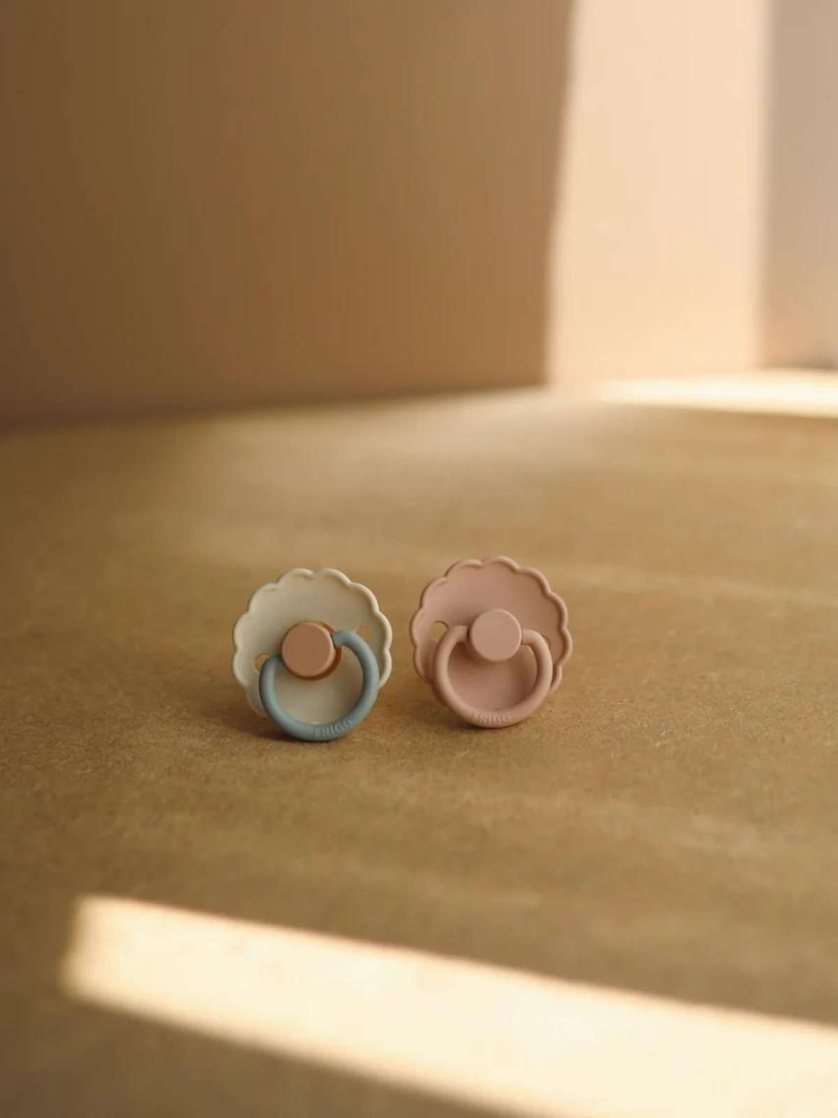 Two pastel-colored pacifiers with scalloped edges on a textured surface, illuminated by soft natural light.