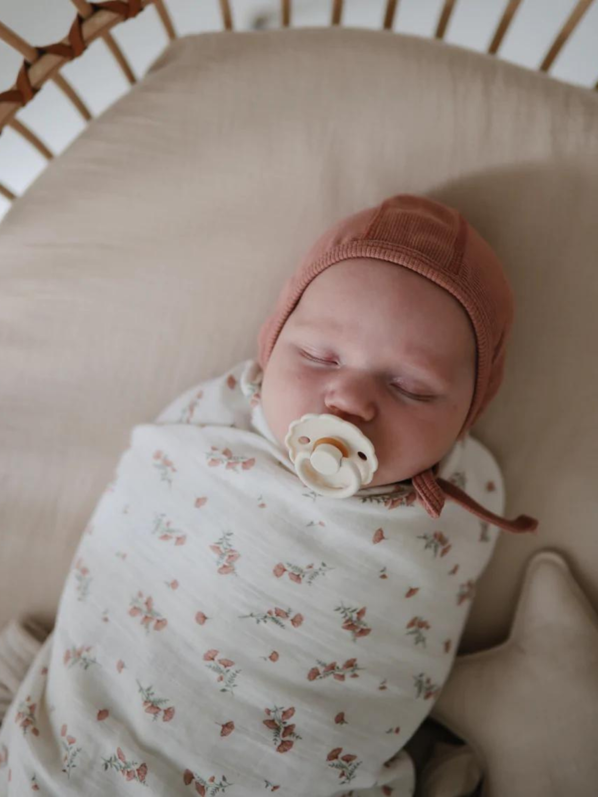 Sleeping baby swaddled in floral-patterned fabric, wearing a brown cap, with a pacifier in a crib.
